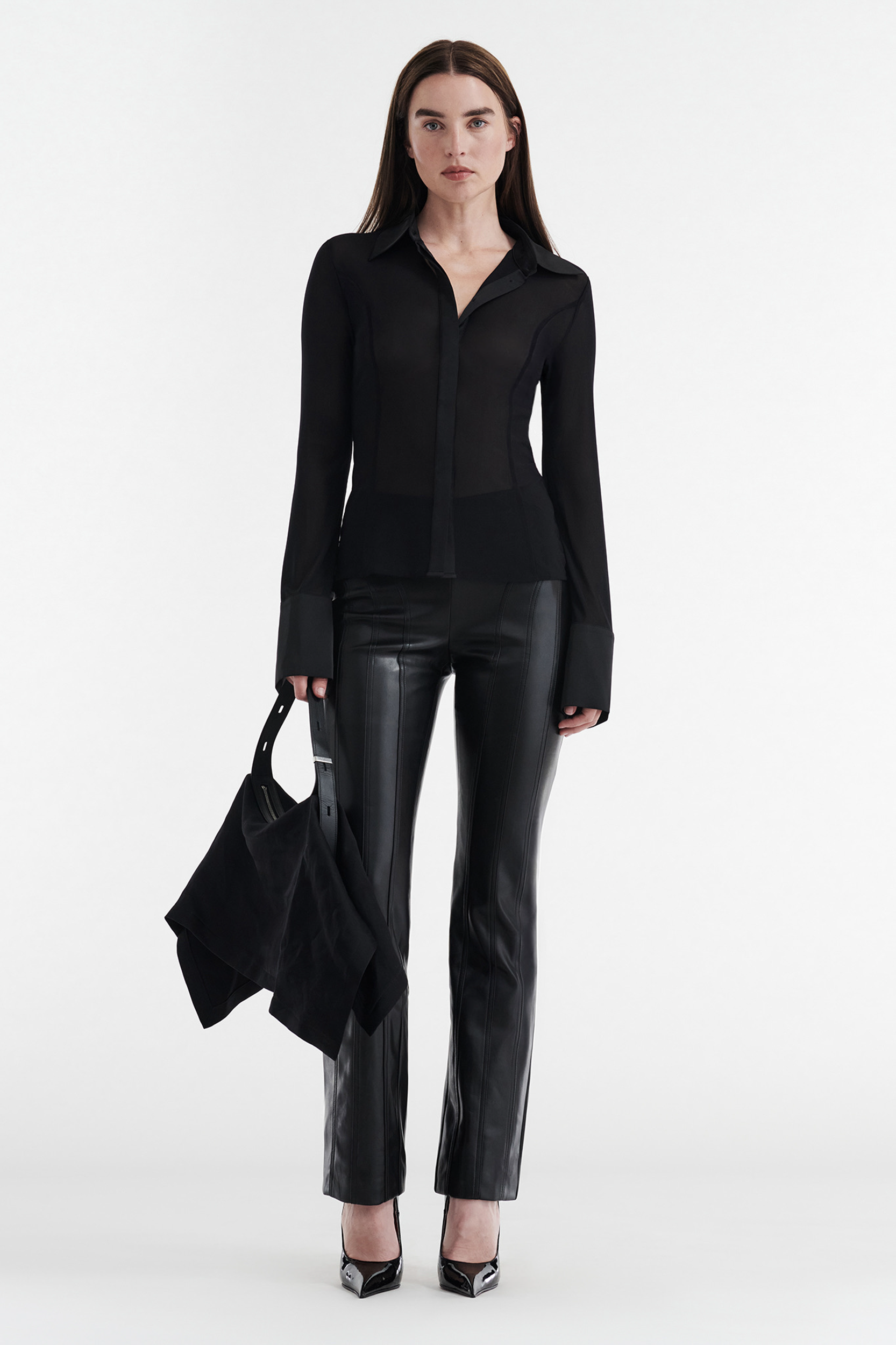 A woman with long straight brown hair and a neutral expression stands against a white background wearing CDP's ALYA FAUX LEATHER MID RISE PANT in black, a sheer black blouse, black heels, and holding a black bag.