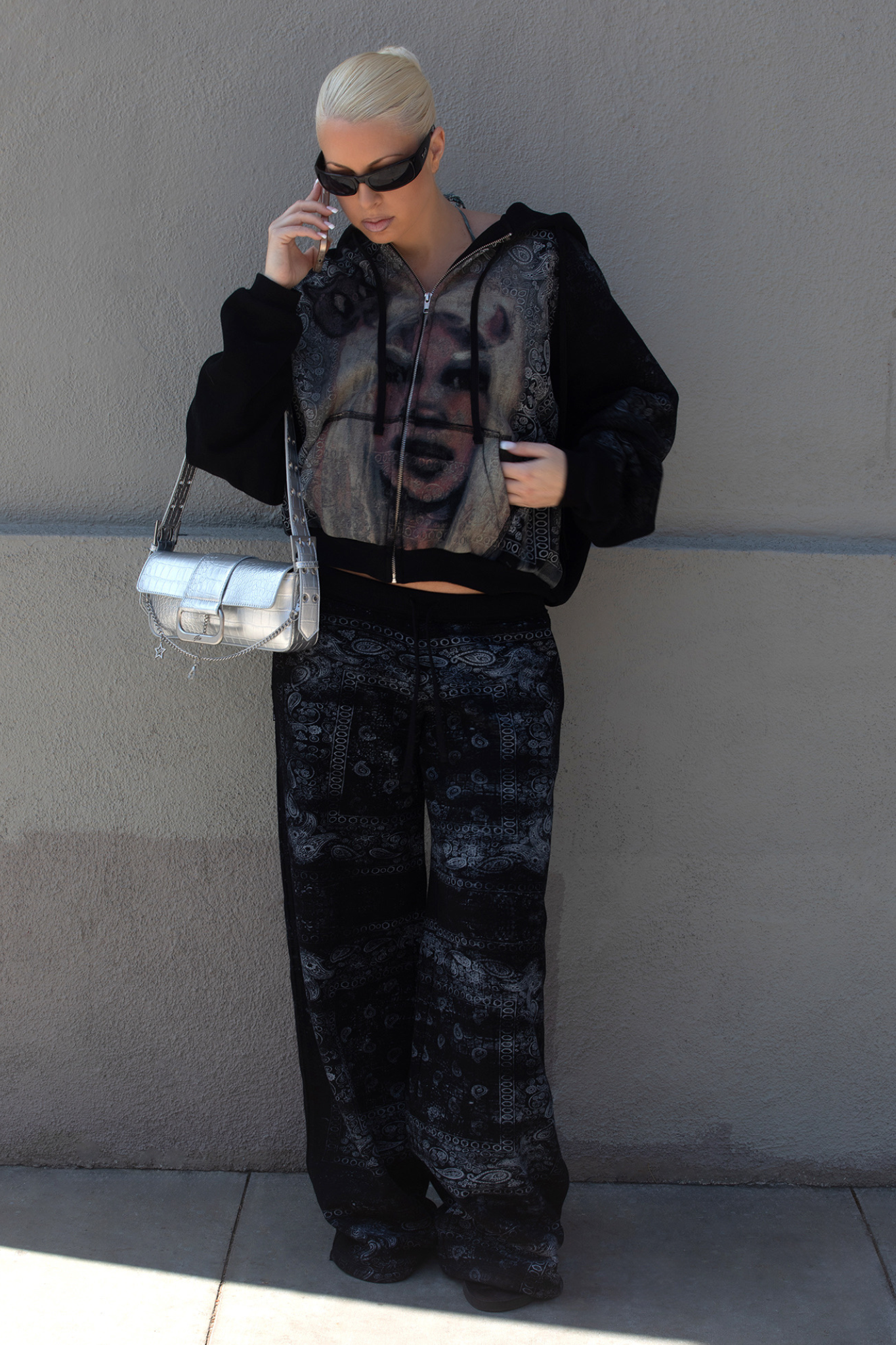 Wearing I.AM.GIA’s T-REX ZIP THRU HOODIE in Black Bandana, a person in dark sunglasses and patterned black pants stands against a wall, holding a silver handbag and talking on a phone.