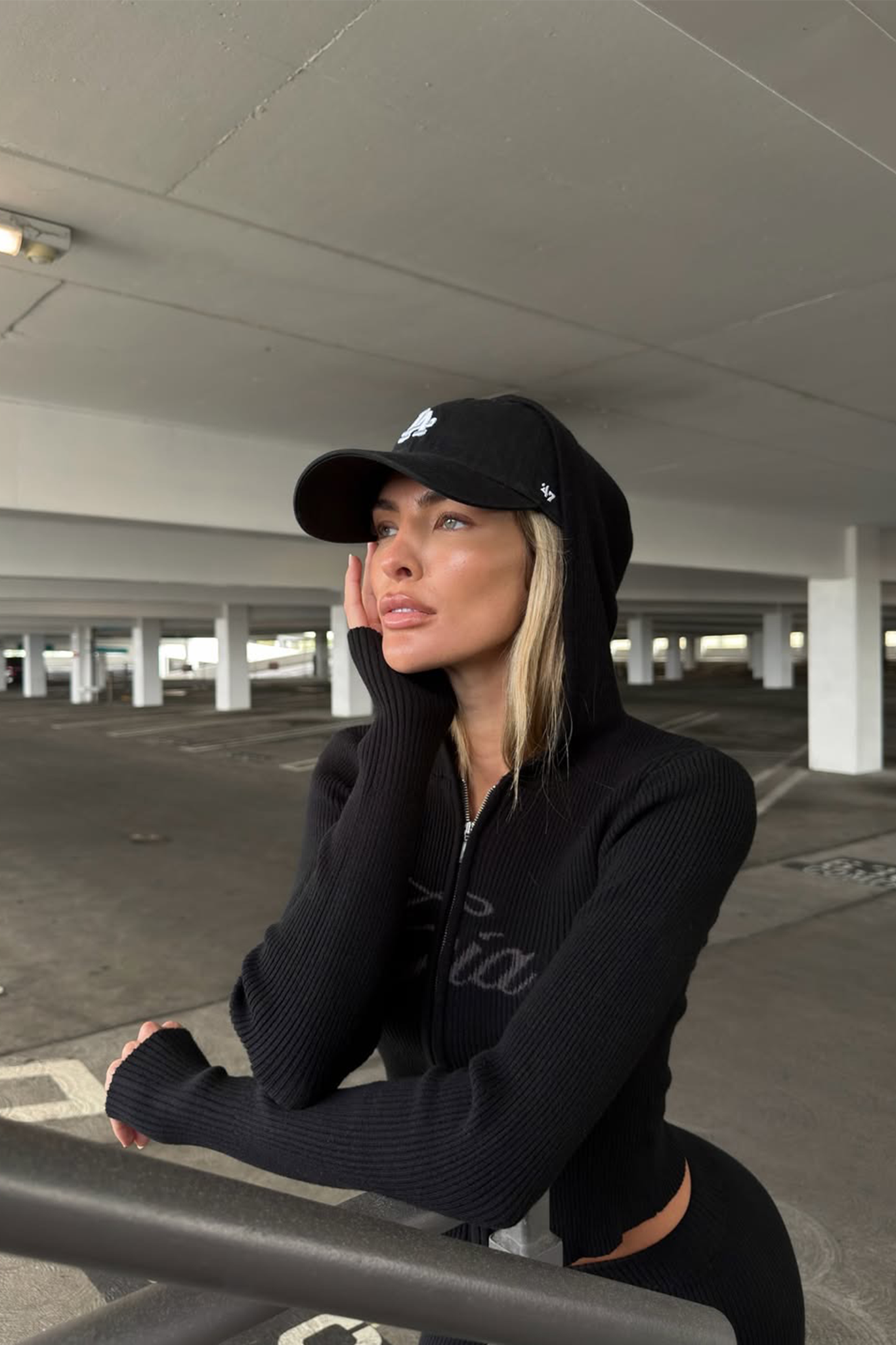 A woman in the I.AM.GIA BLARE KNIT HOODIE - BLACK and a black cap leans on a metal rail in an empty parking garage, gazing thoughtfully into the distance with her hand on her face.