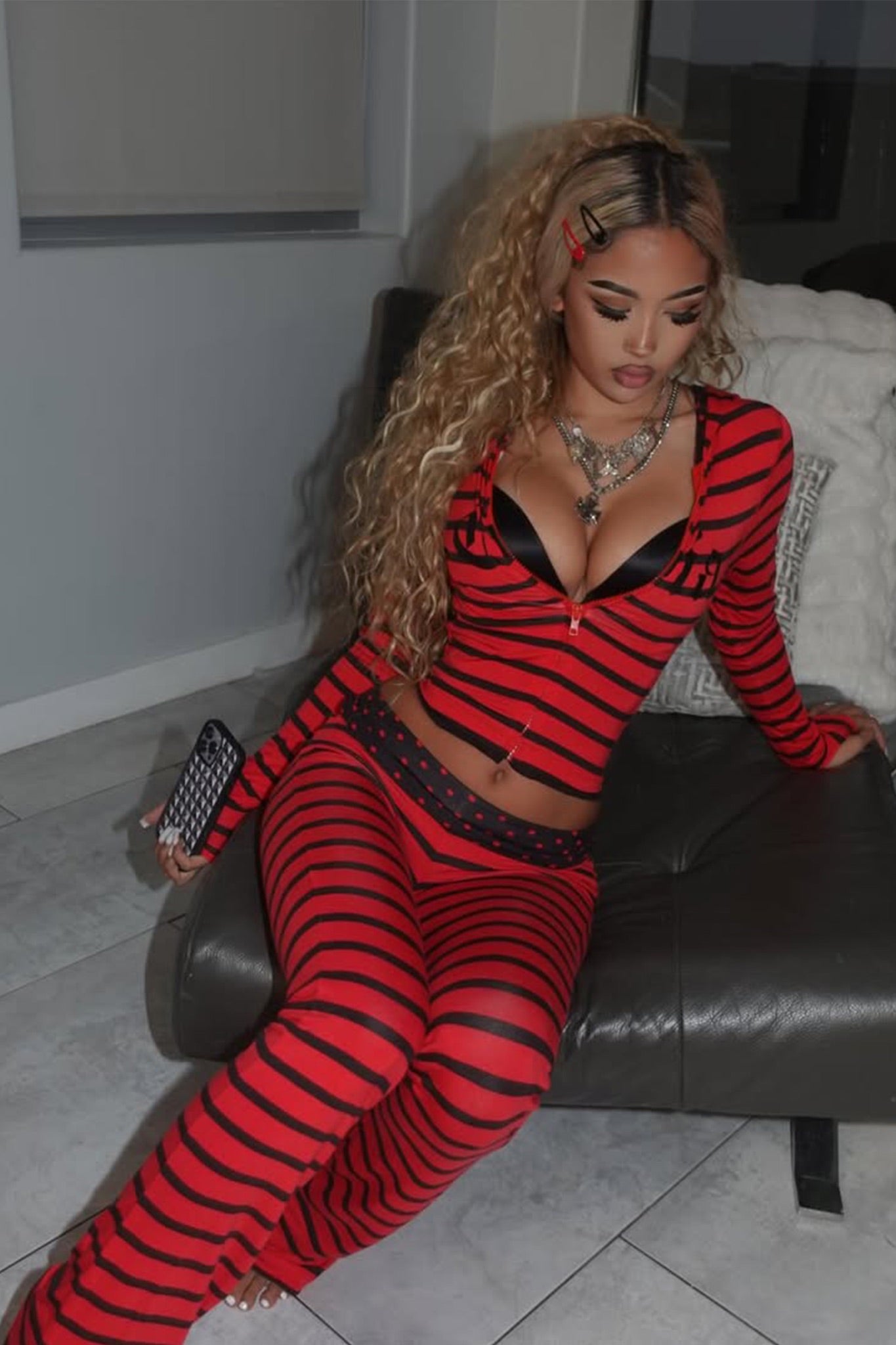 A woman with long curly hair sits on a gray couch, wearing the I.AM.GIA BLARE FOLDOVER TRACKPANT in red and black stripe, with a black bra visible underneath. She holds a phone and looks down, surrounded by neutral-toned decor.