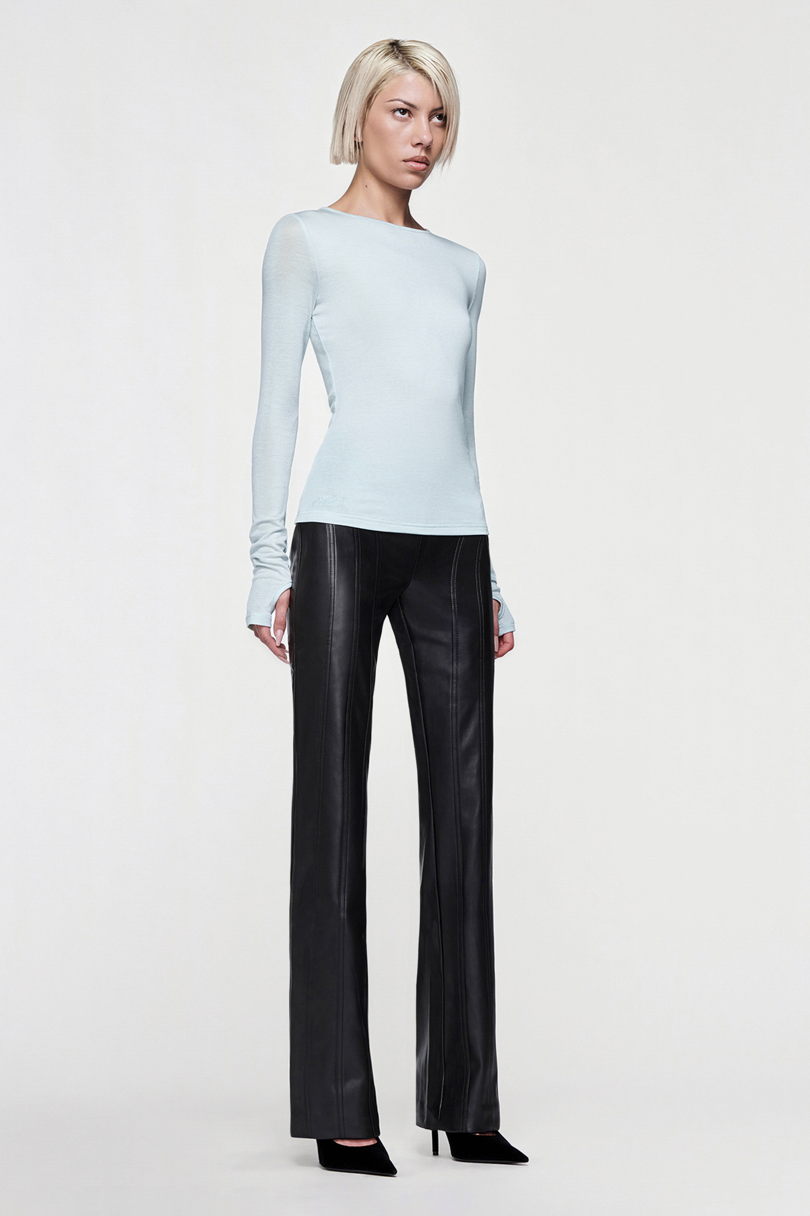 A person with short blond hair wears a fitted CDP CLAUDIA LONG SLEEVE BASIC TOP in eggshell blue, black straight-leg leather pants, and black pointed-toe heels while standing against a plain white background.