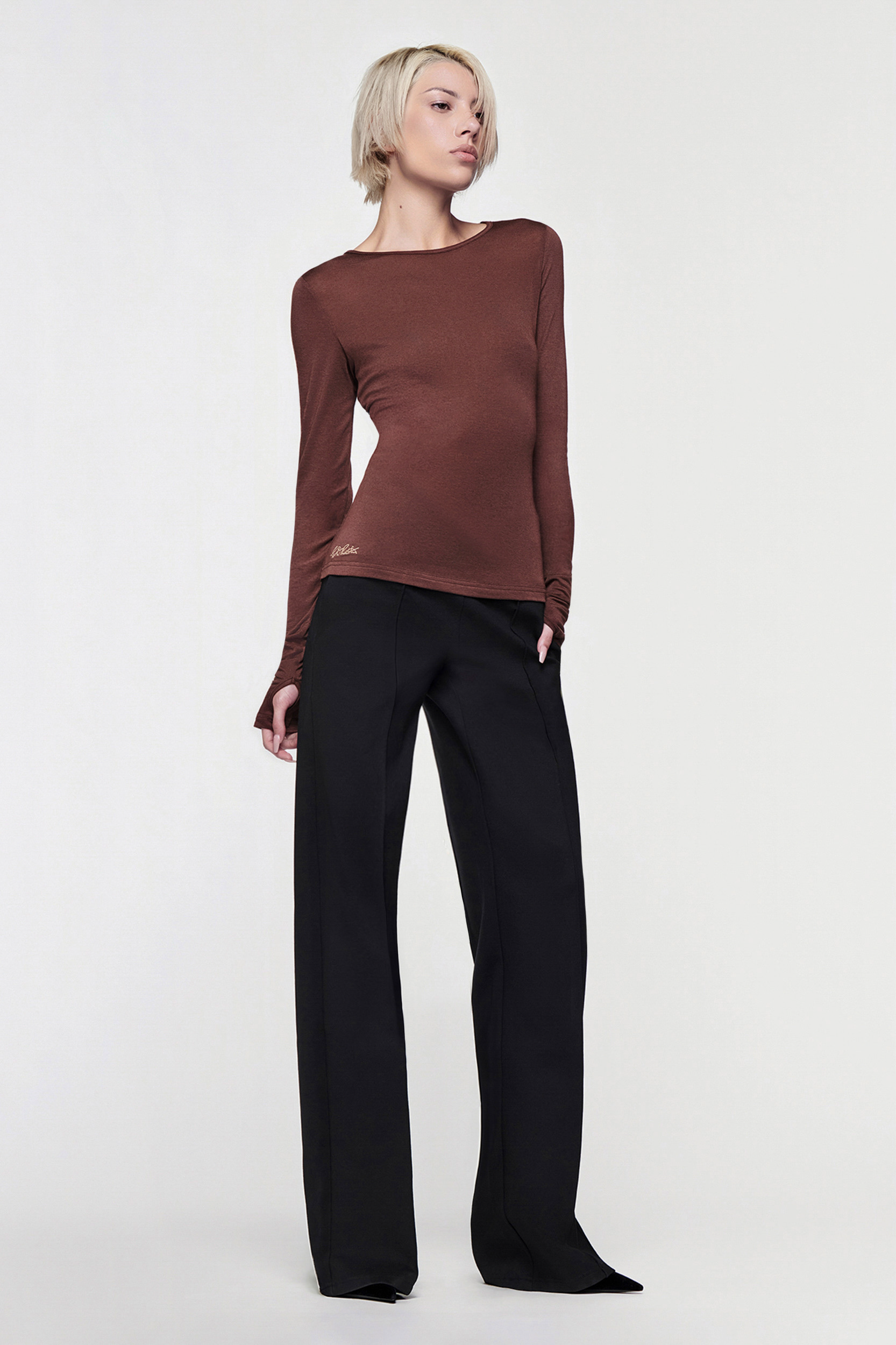 A person with short blond hair models the CDP CLAUDIA LONG SLEEVE BASIC TOP in Chocolate with wide-leg black pants, standing confidently against a plain white background.