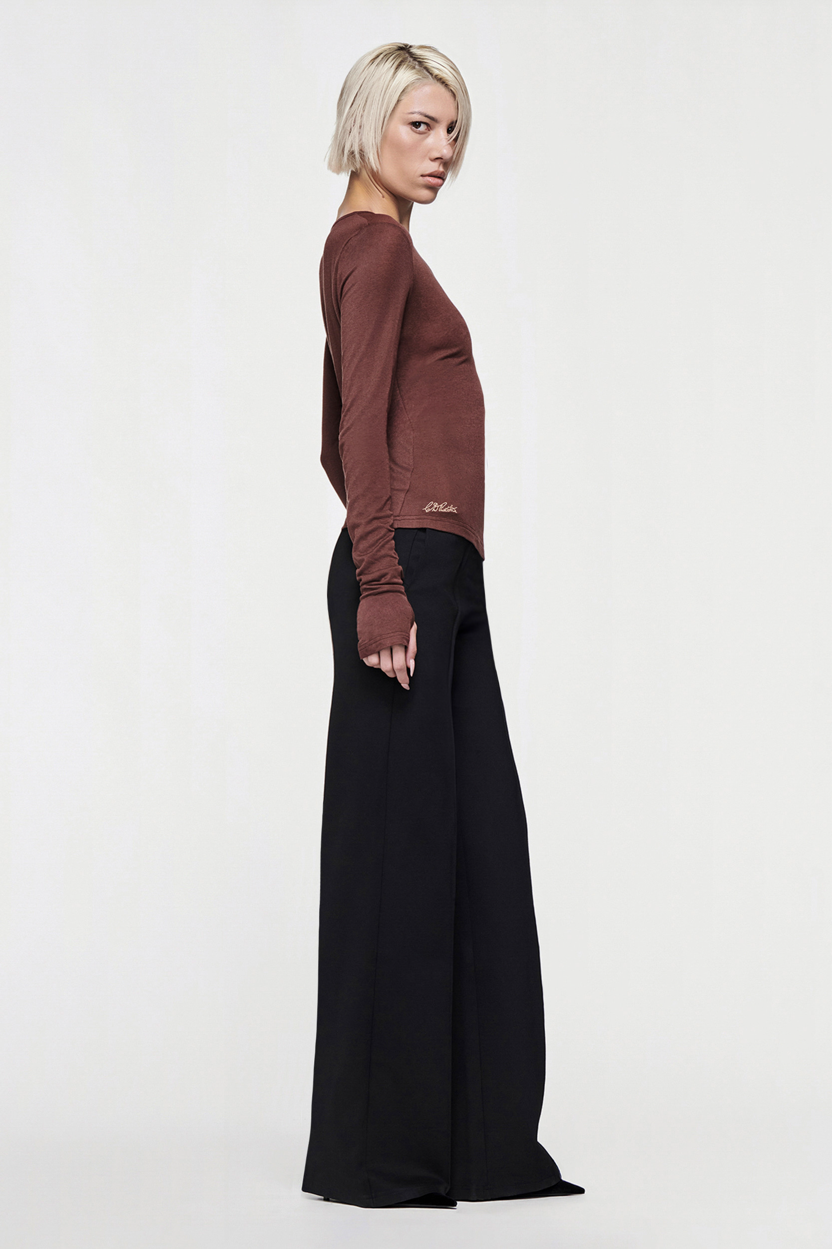A person with short blond hair stands in profile, wearing the CDP CLAUDIA LONG SLEEVE BASIC TOP in Chocolate and high-waisted, wide-leg black pants against a plain light background.