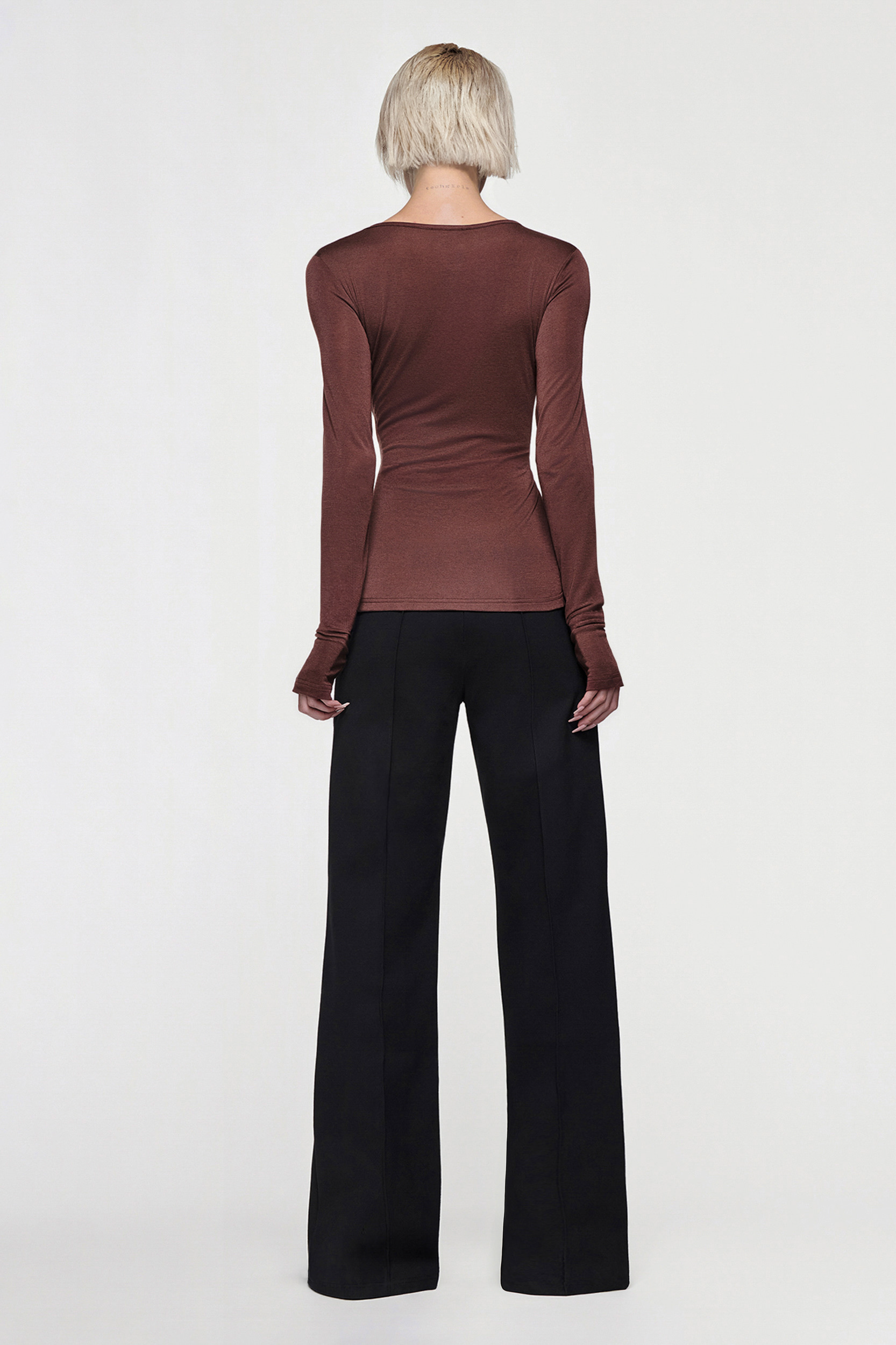 A person with short blonde hair faces away, wearing the CDP CLAUDIA LONG SLEEVE BASIC TOP - CHOCOLATE in a fitted silhouette and high-waisted black wide-leg pants against a plain white background.