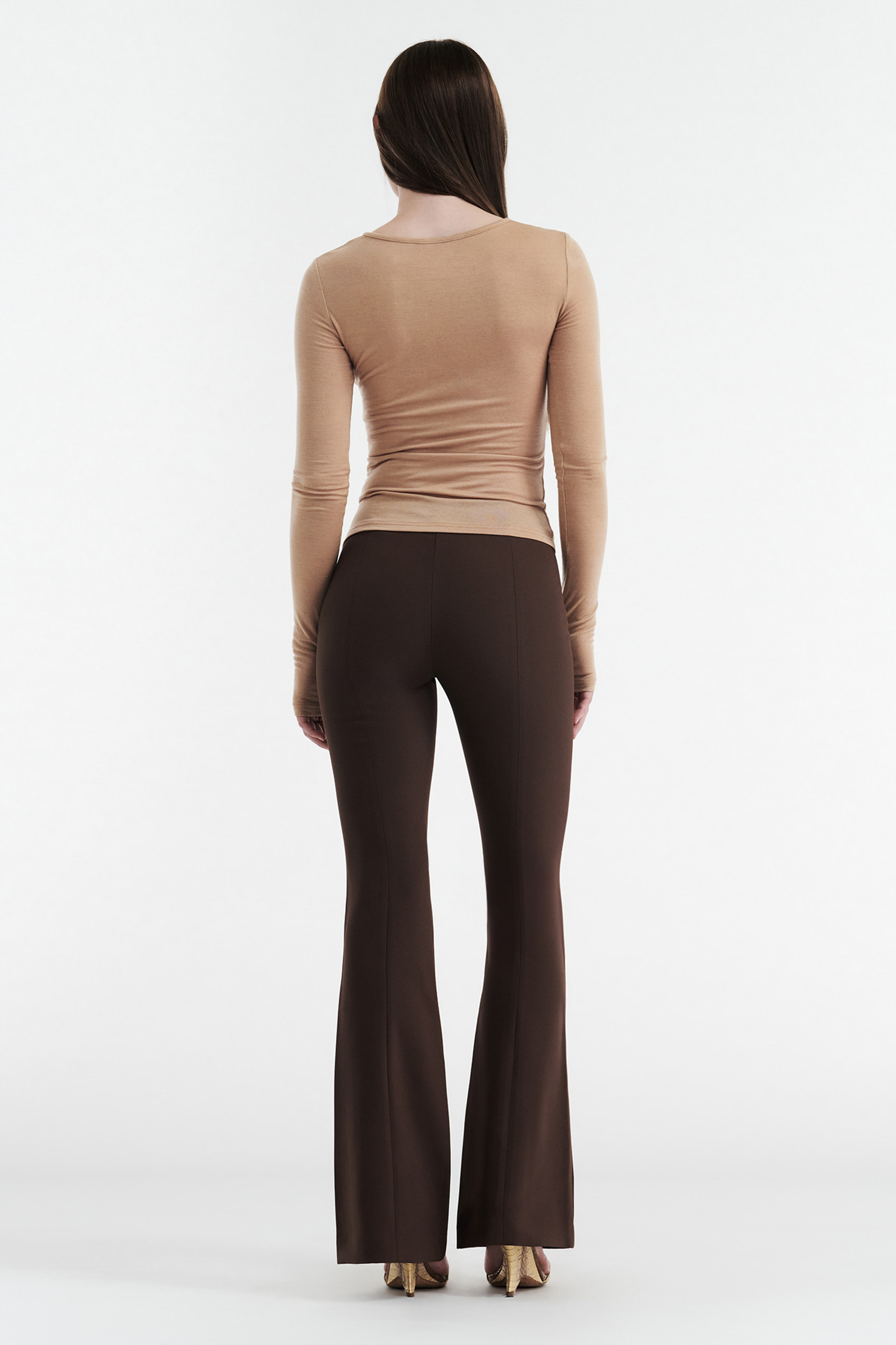 A person with long, straight brown hair stands facing away, wearing the CDP CLAUDIA LONG SLEEVE BASIC TOP in tan, paired with dark brown flared pants and beige shoes, against a plain white background.