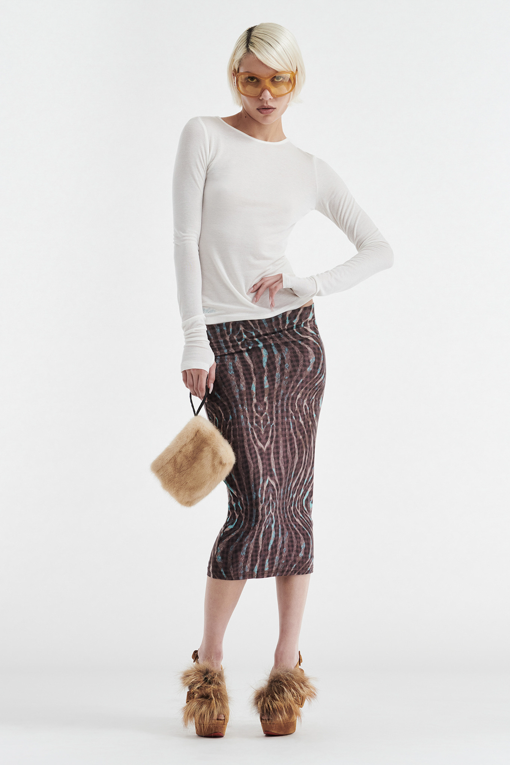 A woman with short platinum blonde hair wears yellow sunglasses, a CDP CLAUDIA LONG SLEEVE BASIC TOP in white, a patterned brown midi skirt, furry brown platform heels, and holds a small fuzzy beige handbag. She poses against a white background.