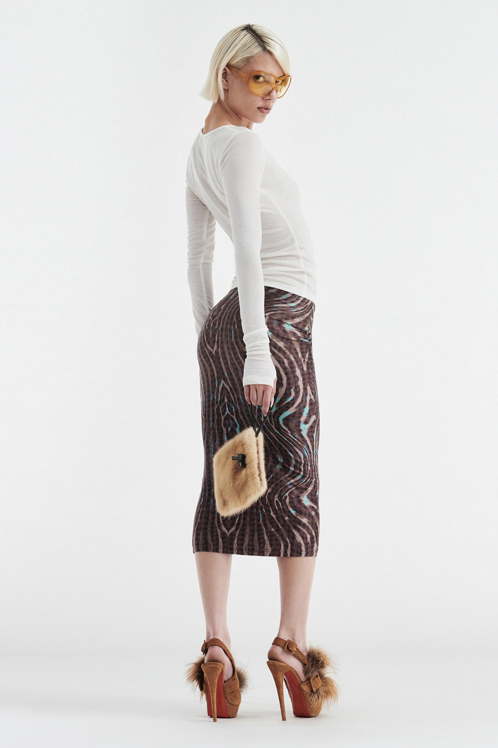 A person with short blonde hair and yellow sunglasses wears a CDP CLAUDIA LONG SLEEVE BASIC TOP in white, paired with a brown animal-print skirt, fur-accented wooden platform heels, and a small fur purse, posed against a white background.