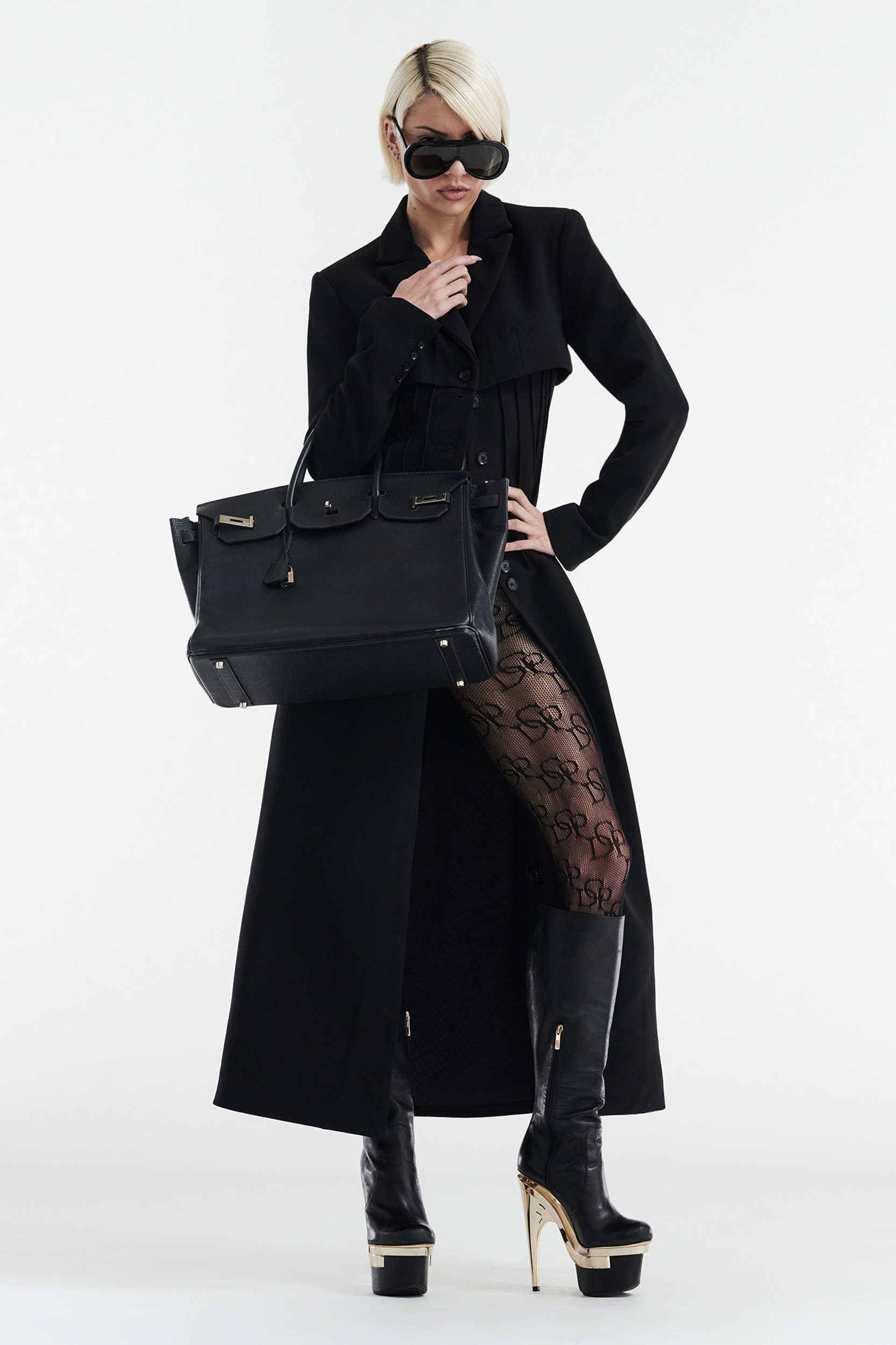 Wearing the CELINE BUTTON UP TRENCH COAT in black by CDP, a person pairs it with patterned tights, platform boots with gold heels, large sunglasses, and carries a big black handbag against a plain white background.