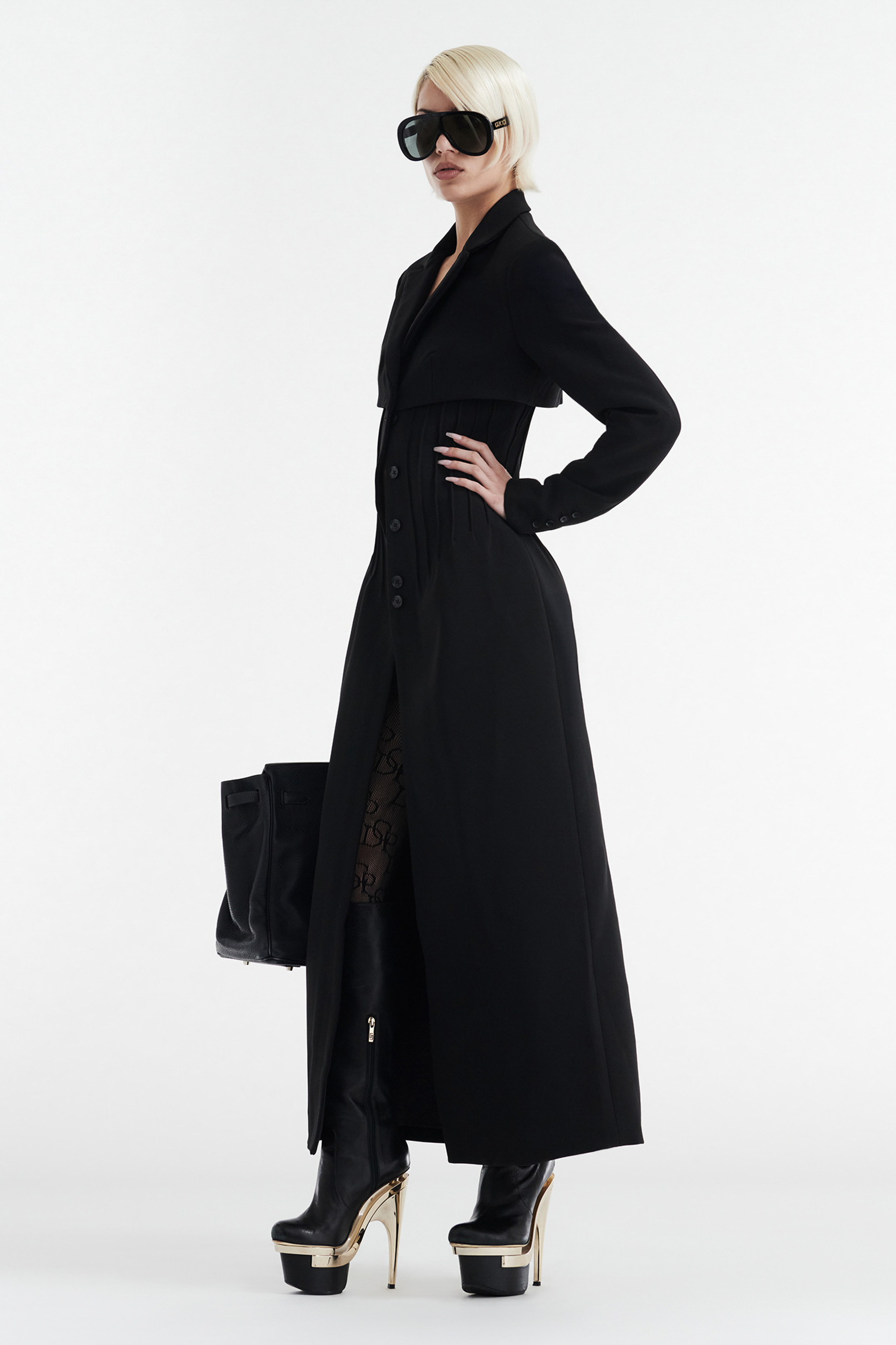 A person with short blond hair and large black sunglasses stands in profile, wearing a CDP CELINE BUTTON UP TRENCH COAT - BLACK, patterned stockings, platform boots with gold heels, and holding a large black bag.