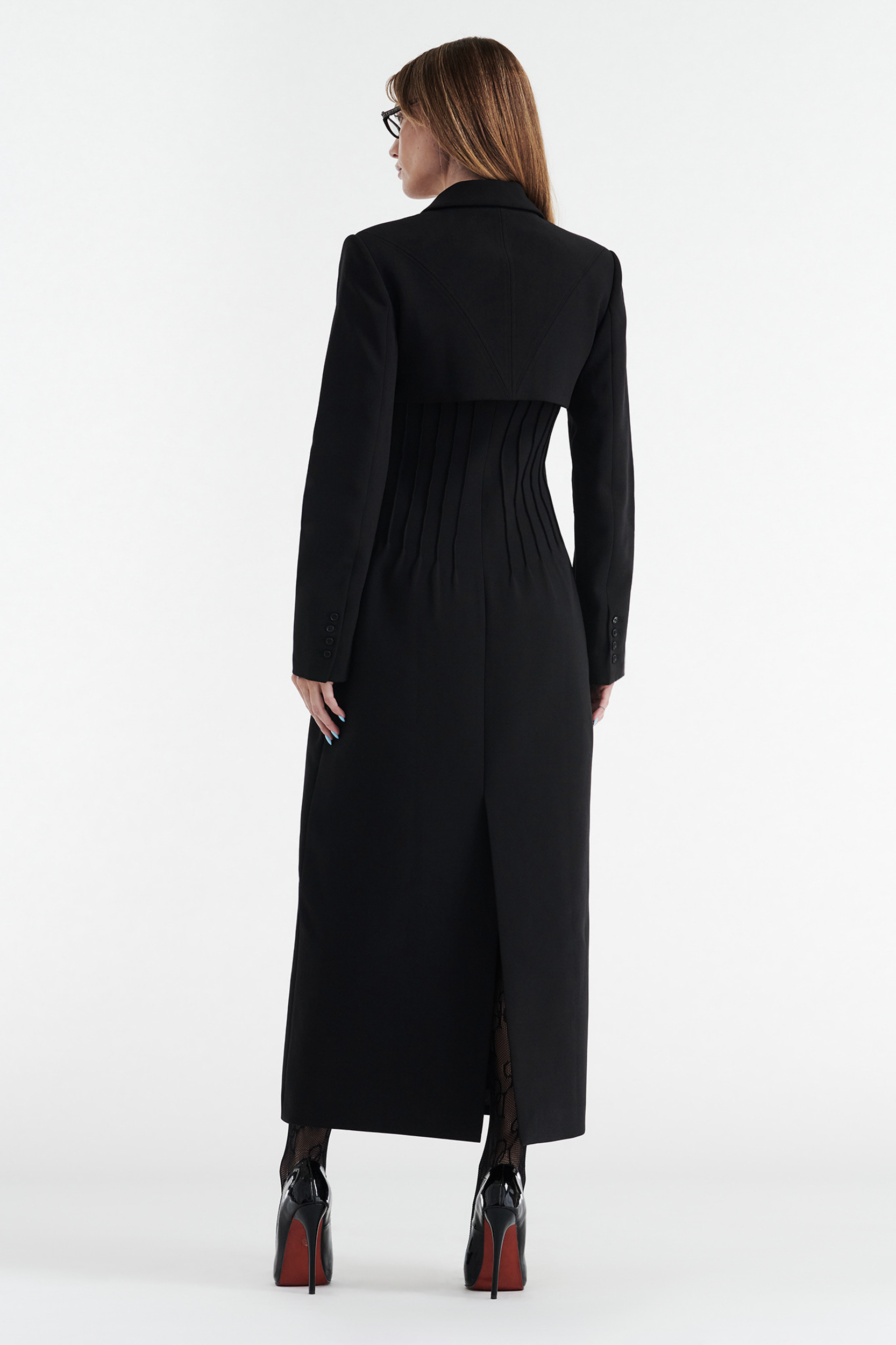 A woman with straight brown hair and glasses faces away, modeling the CDP CELINE BUTTON UP TRENCH COAT - BLACK with vertical pleats and a tailored fit, paired with black high heels against a plain white background.