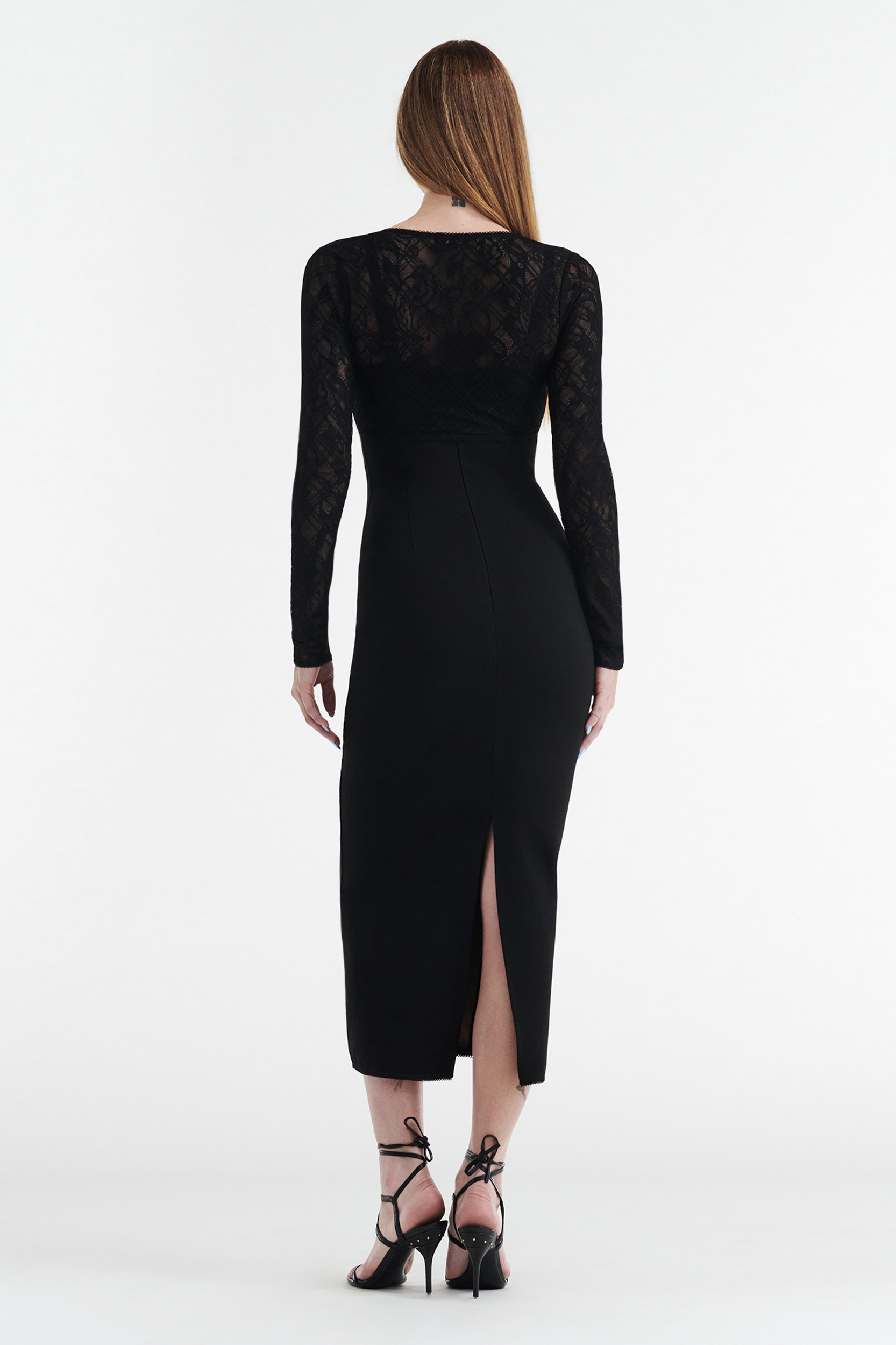 A woman with long straight hair faces away, wearing the CDP MARGOT Two Piece Fitted Lace Midi Dress in black, featuring sheer lace sleeves and back, a mid-calf hem with back slit, and paired with black strappy high-heeled sandals.