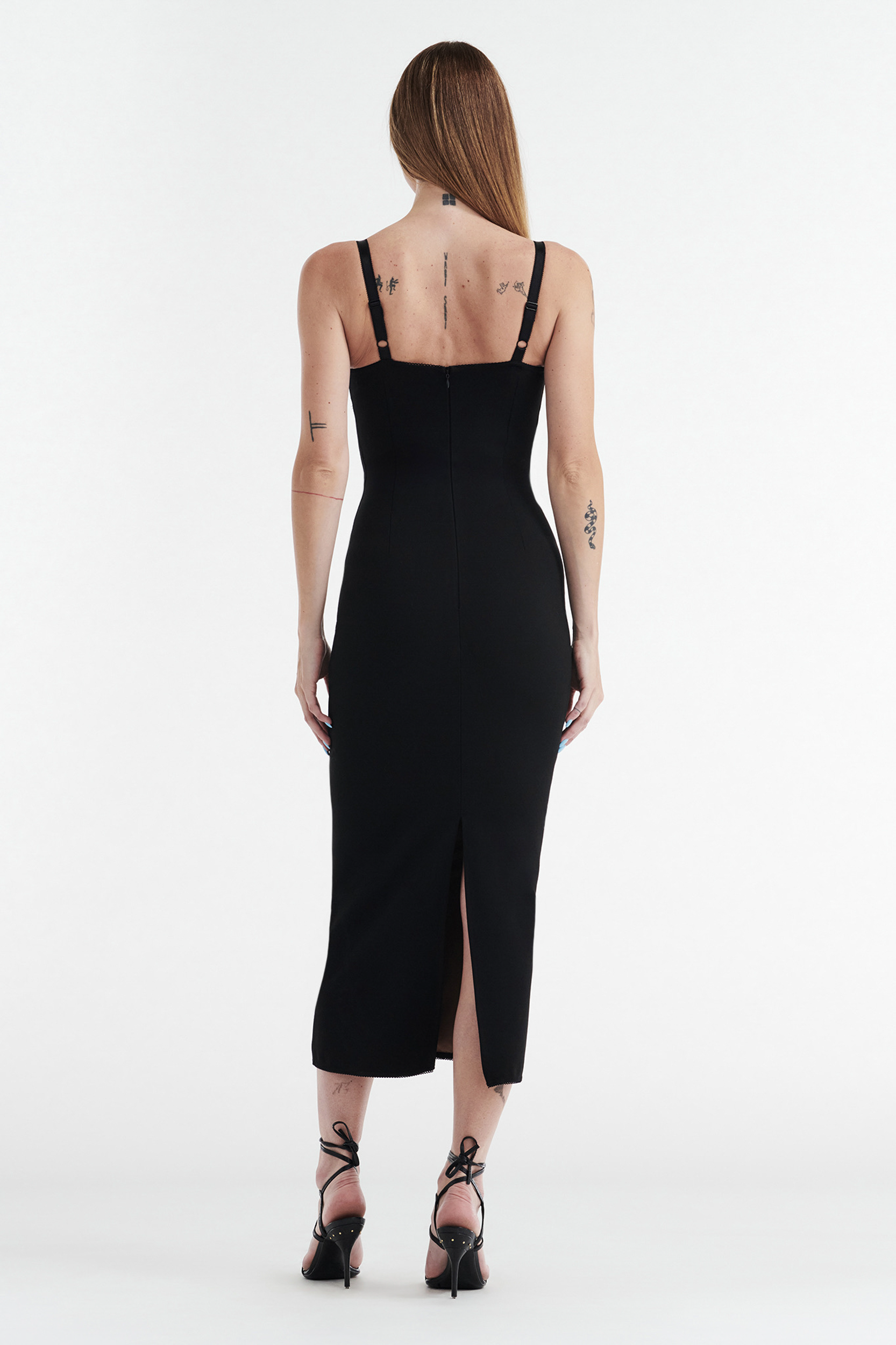 A woman with long straight hair faces away, wearing the CDP MARGOT Two Piece Fitted Lace Midi Dress in black with thin straps and a back slit, paired with black high-heeled sandals. Her arms and upper back feature tattoos against a white background.