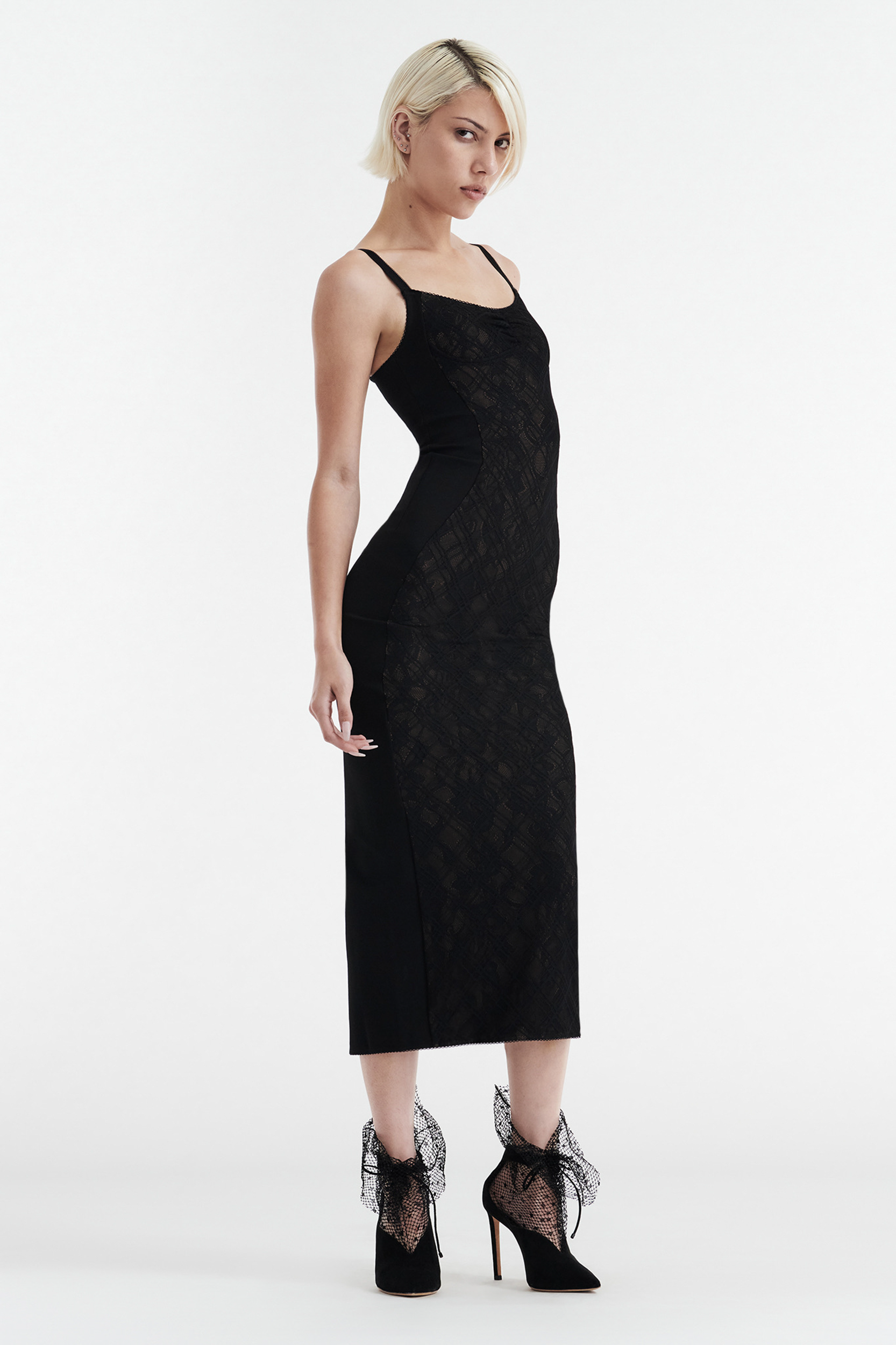 A woman with short blonde hair models the CDP MARGOT TWO PIECE FITTED LACE MIDI DRESS in black. She stands sideways on a white background, wearing black high heels with mesh bows and thin straps.