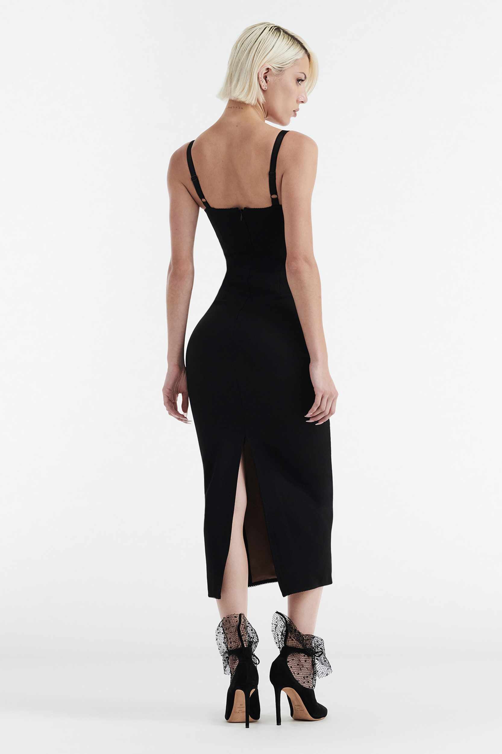 A woman with short blonde hair faces away in the CDP MARGOT TWO PIECE FITTED LACE MIDI DRESS - BLACK, featuring a back slit. She pairs it with black high heels with mesh ankle details against a plain white background.