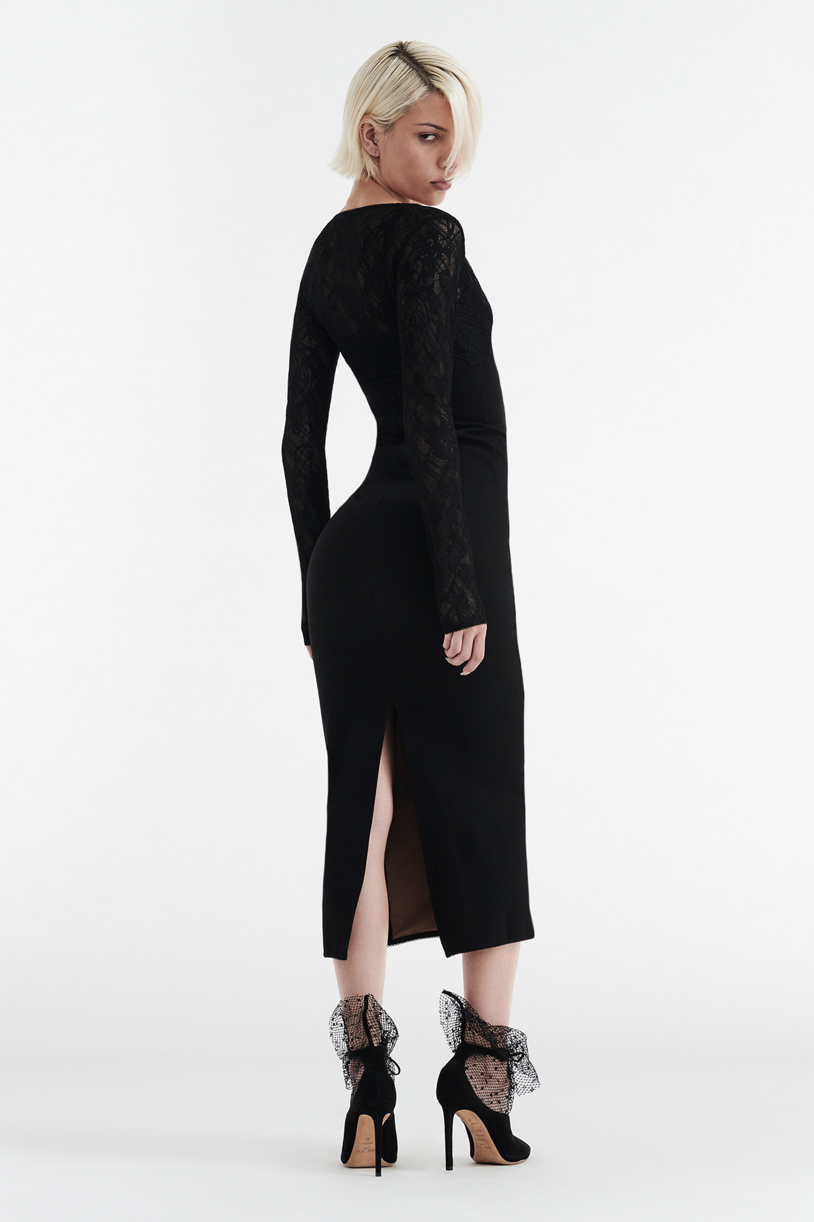 A woman with short blonde hair models the CDP MARGOT Two Piece Fitted Lace Midi Dress in black, featuring a back slit. She pairs it with black high heels with sheer ruffled ankle bows, posing against a plain white background.