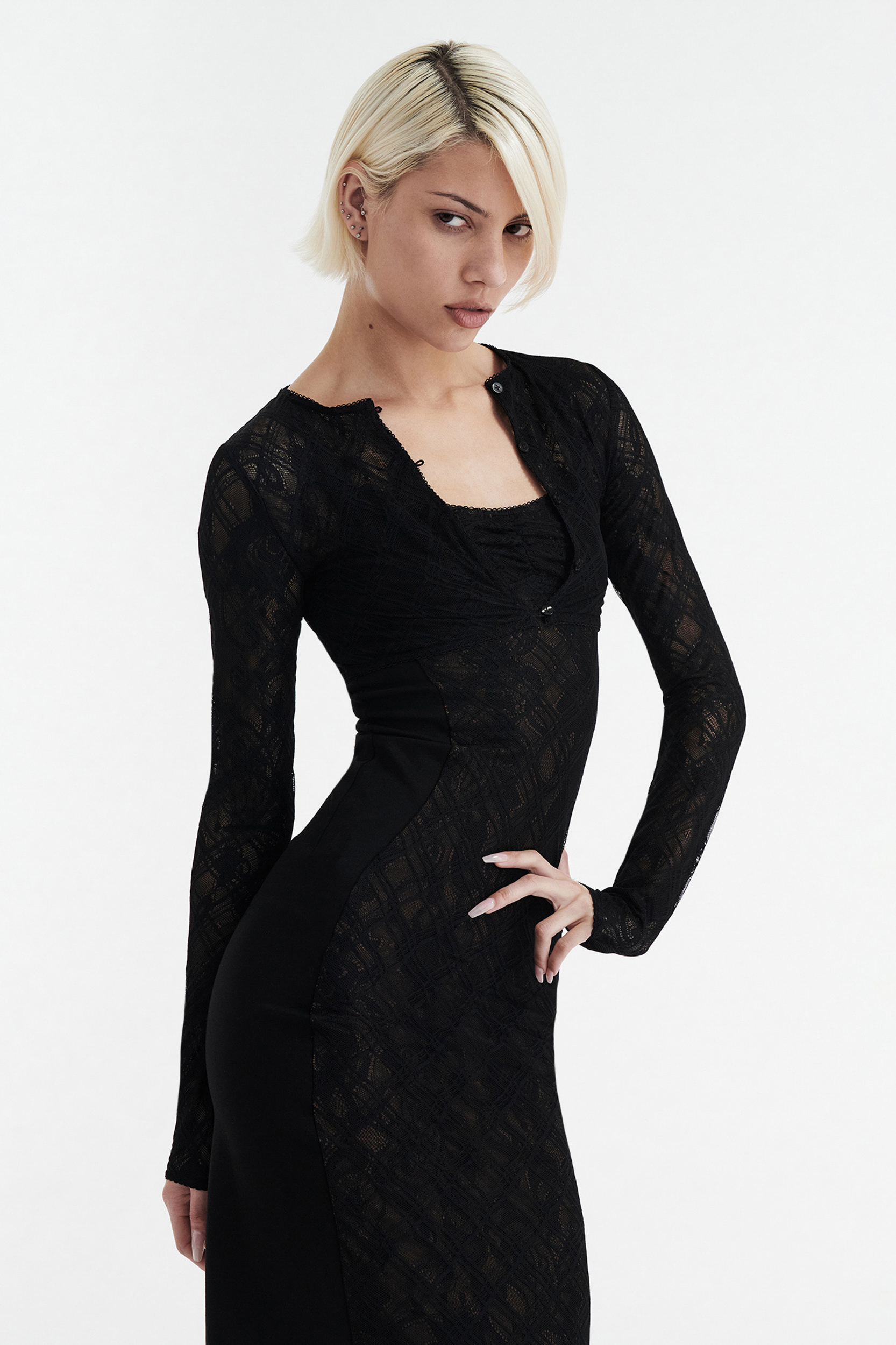 A person with short blonde hair models the CDP MARGOT TWO PIECE FITTED LACE MIDI DRESS in black, posing with one hand on their hip against a plain white background.