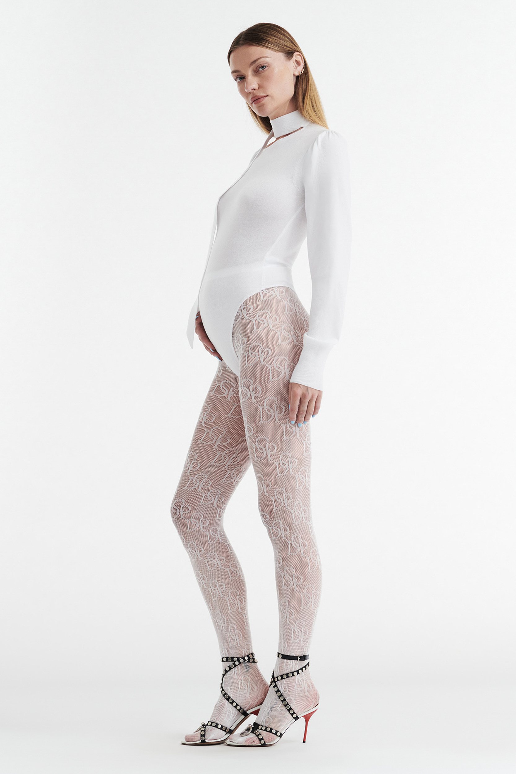 A woman stands in profile against a white background wearing the CDP VIVIENNE LONG SLEEVE KNIT BODYSUIT in white, sheer patterned tights with a repeating white design, and open-toed high-heeled sandals with ankle straps.