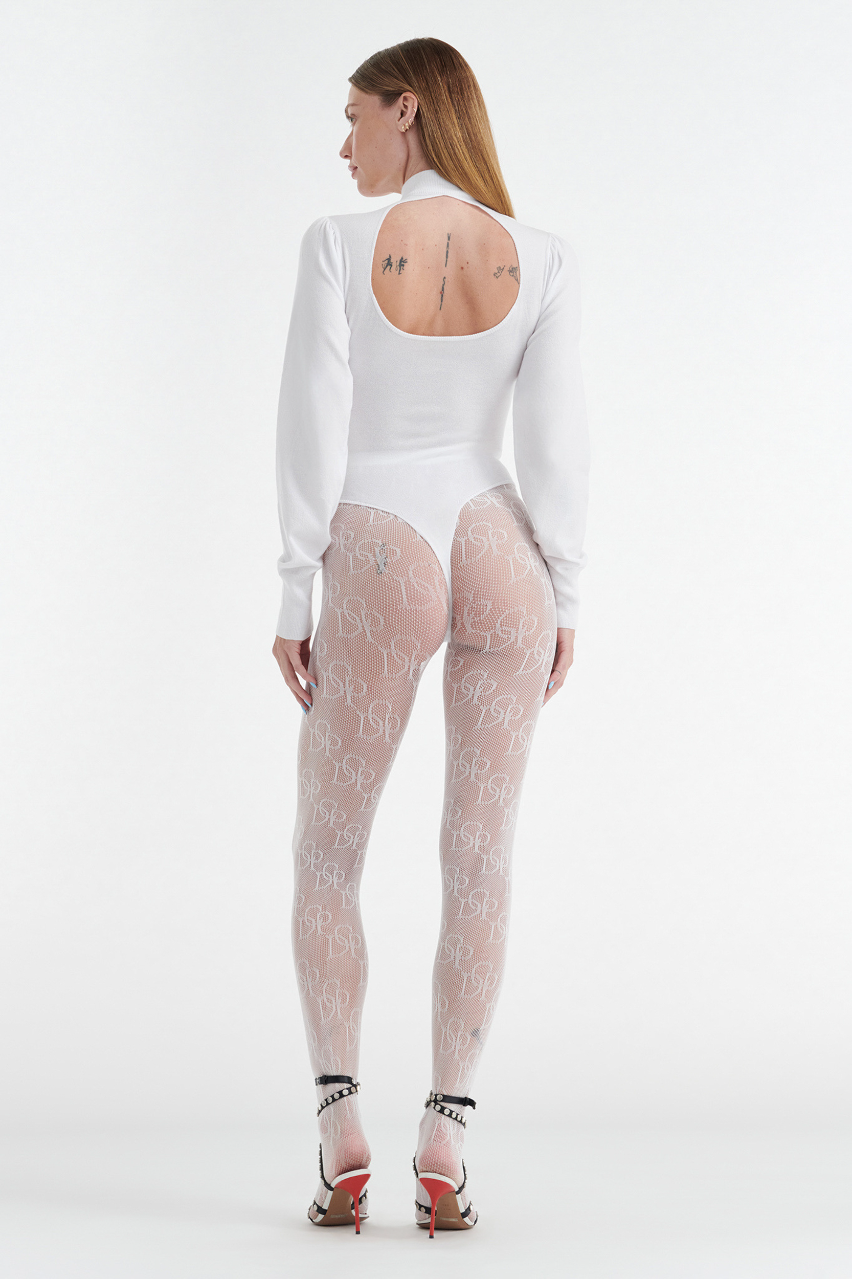 A woman faces away, wearing the CDP VIVIENNE LONG SLEEVE KNIT BODYSUIT in white with sheer logo tights and embellished heels. Her straight hair falls down, exposing tattoos on her upper back.