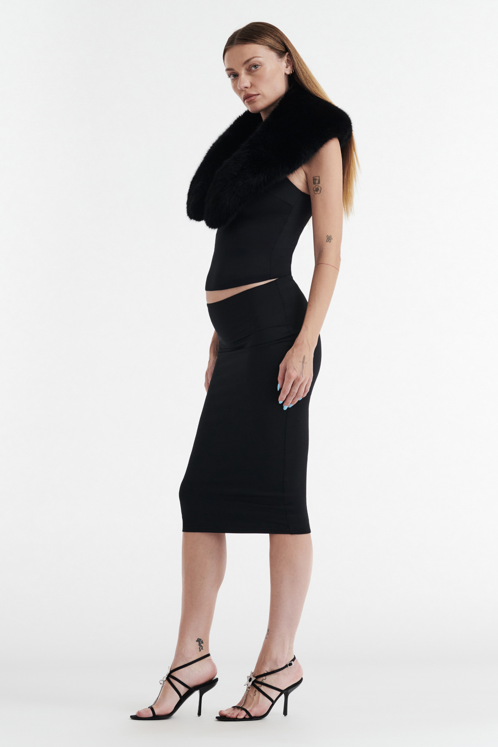 A woman in a fitted black dress with the CDP SHIRLEY FAUX FUR COLLAR - BLACK stands in profile against a white background, wearing strappy heels, showing arm and ankle tattoos, and has straight, loose hair.