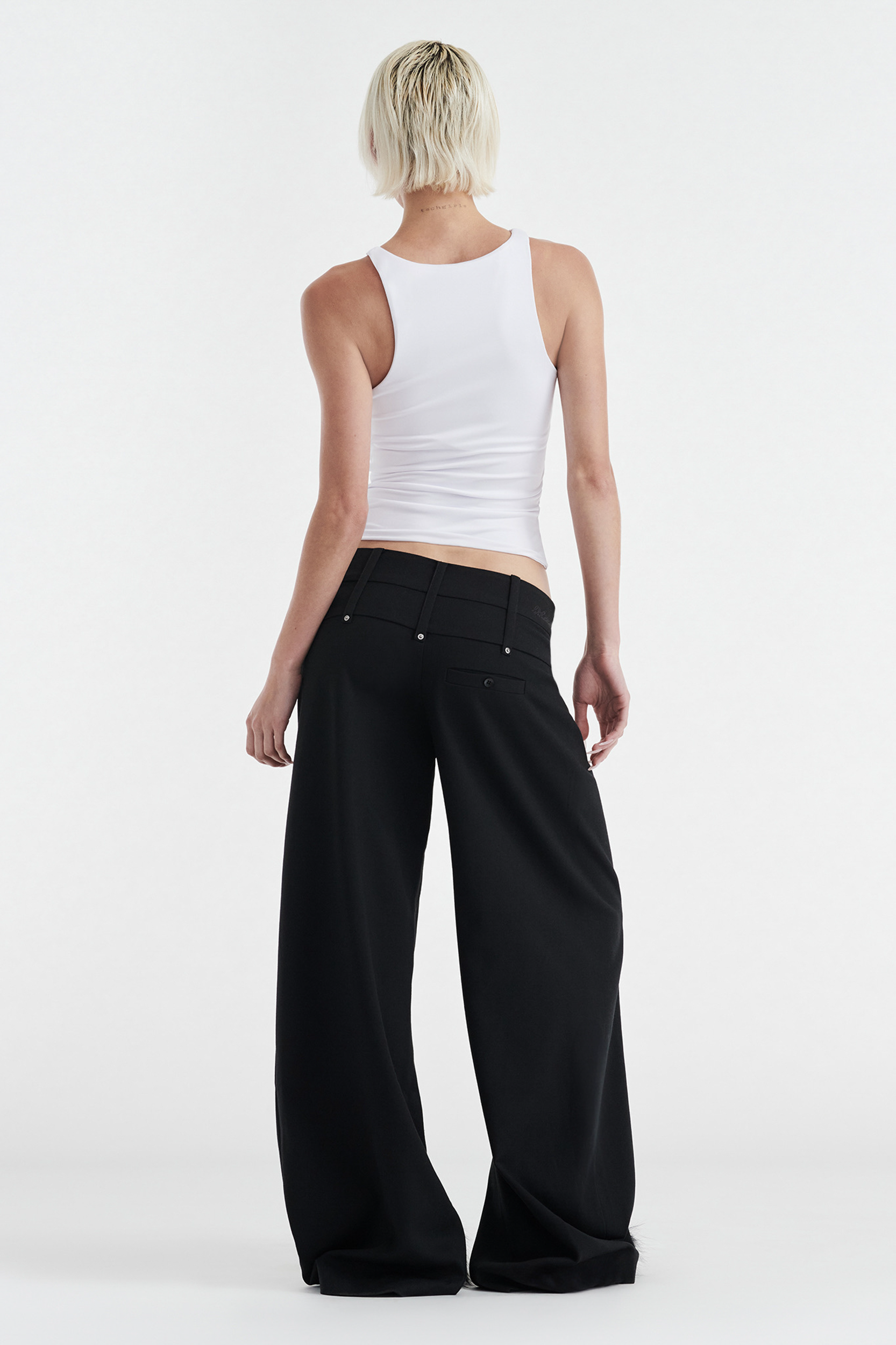 A person with short, light blonde hair faces away against a white background, wearing a fitted white tank top and CDP's CHARLOTTE WIDE LEG PLEAT PANT in black.