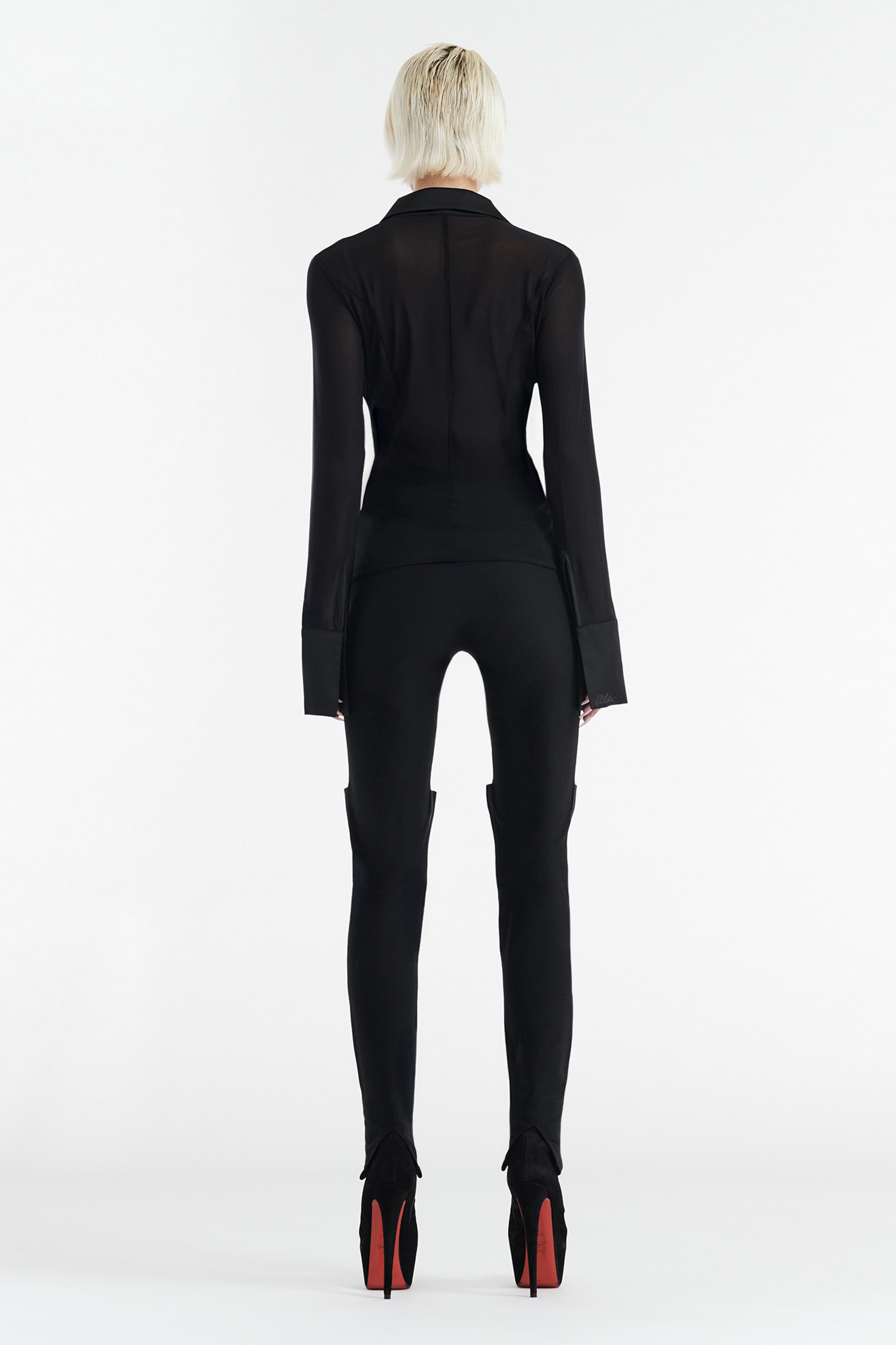 A person with short platinum-blonde hair is seen from behind, wearing a fitted sheer black top and CDP AIMEE High Waisted Slim Fit Pant in black, styled with high-heeled red-soled shoes against a plain white background.