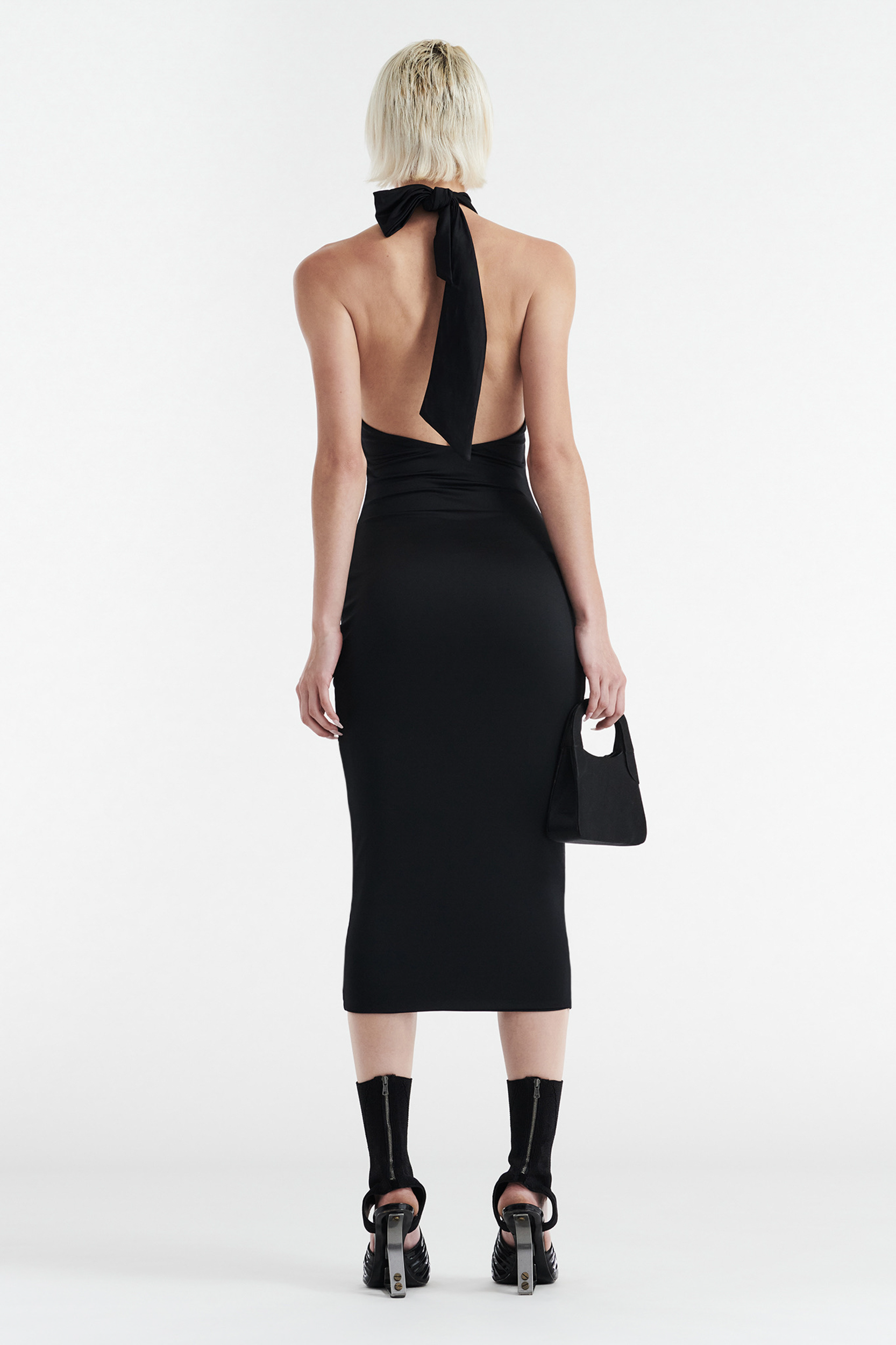A person with short blonde hair wears the CDP CLEO HALTER TIE MIDI DRESS - BLACK, styled with black high-heeled sandals, black ankle socks, and a small black handbag while standing against a plain white background.