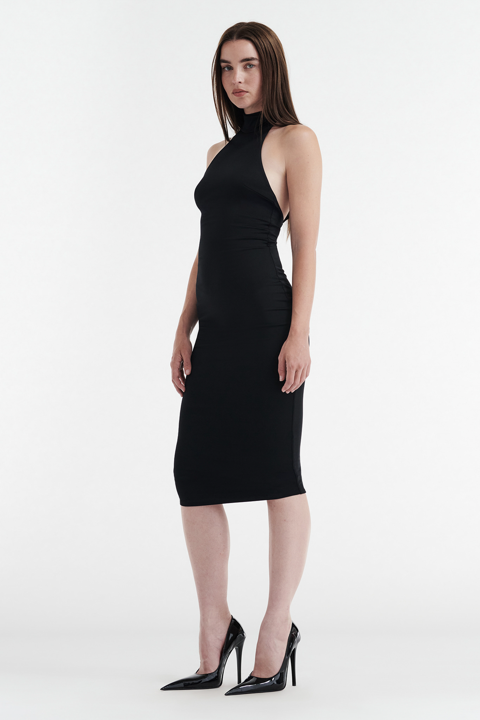 A woman stands against a white background, wearing the CDP CLEO HALTER TIE MIDI DRESS in black with black high heels, looking forward with a neutral expression.