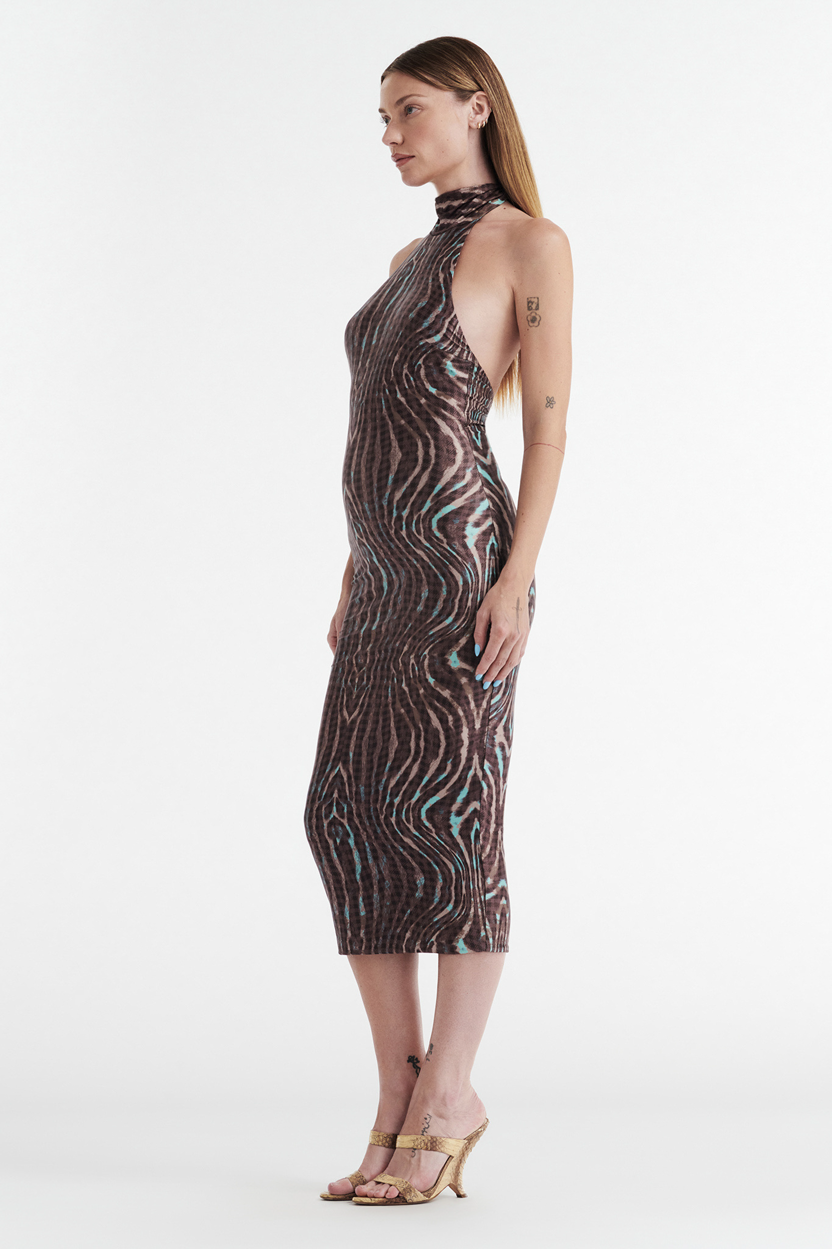 A woman with long hair and visible tattoos models the CDP CLEO HALTER TIE MIDI DRESS - WARPED CHECK, a sleeveless, form-fitting dress featuring an abstract brown, black, and teal pattern. She pairs it with beige high-heeled sandals against a white background.