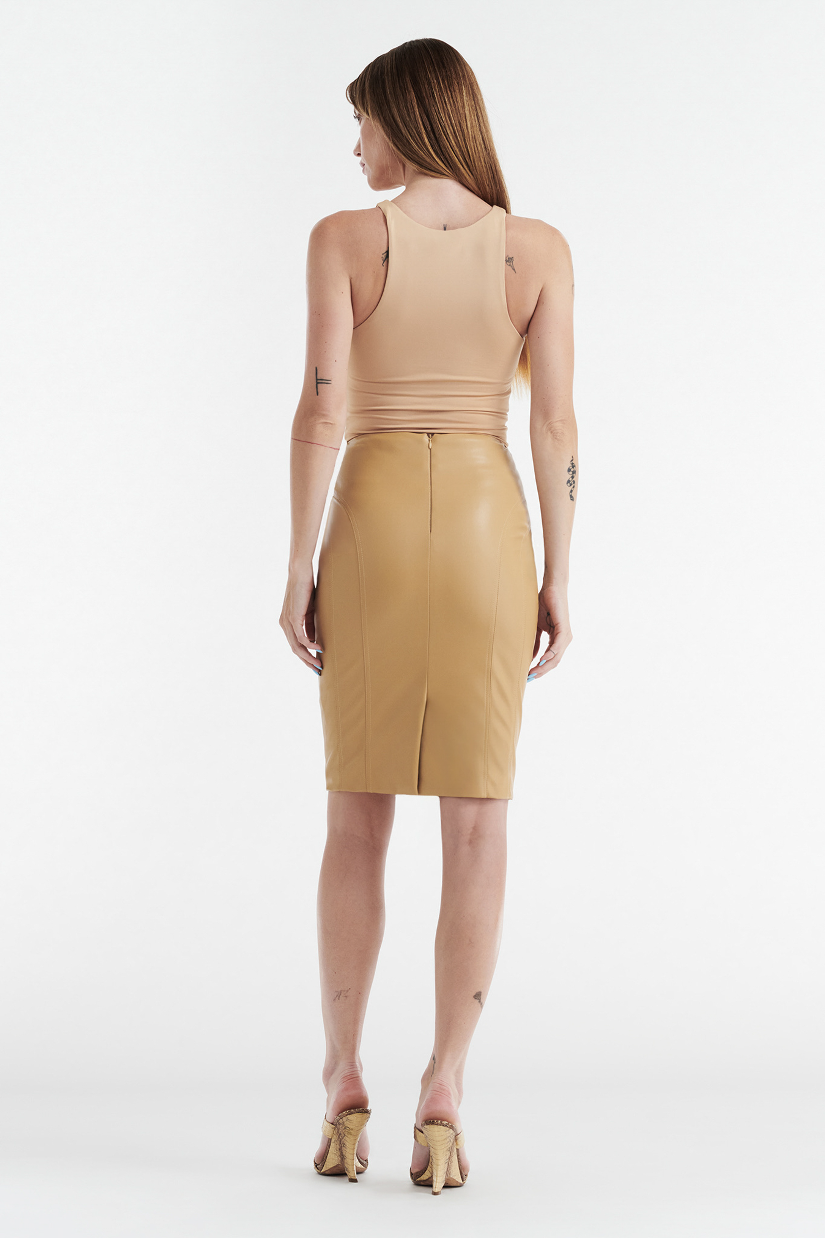 A woman with straight hair and visible arm and ankle tattoos stands facing away against a white background, wearing a fitted beige sleeveless top, the CDP INES FAUX LEATHER PENCIL SKIRT in tan, midi-length, and heeled sandals.