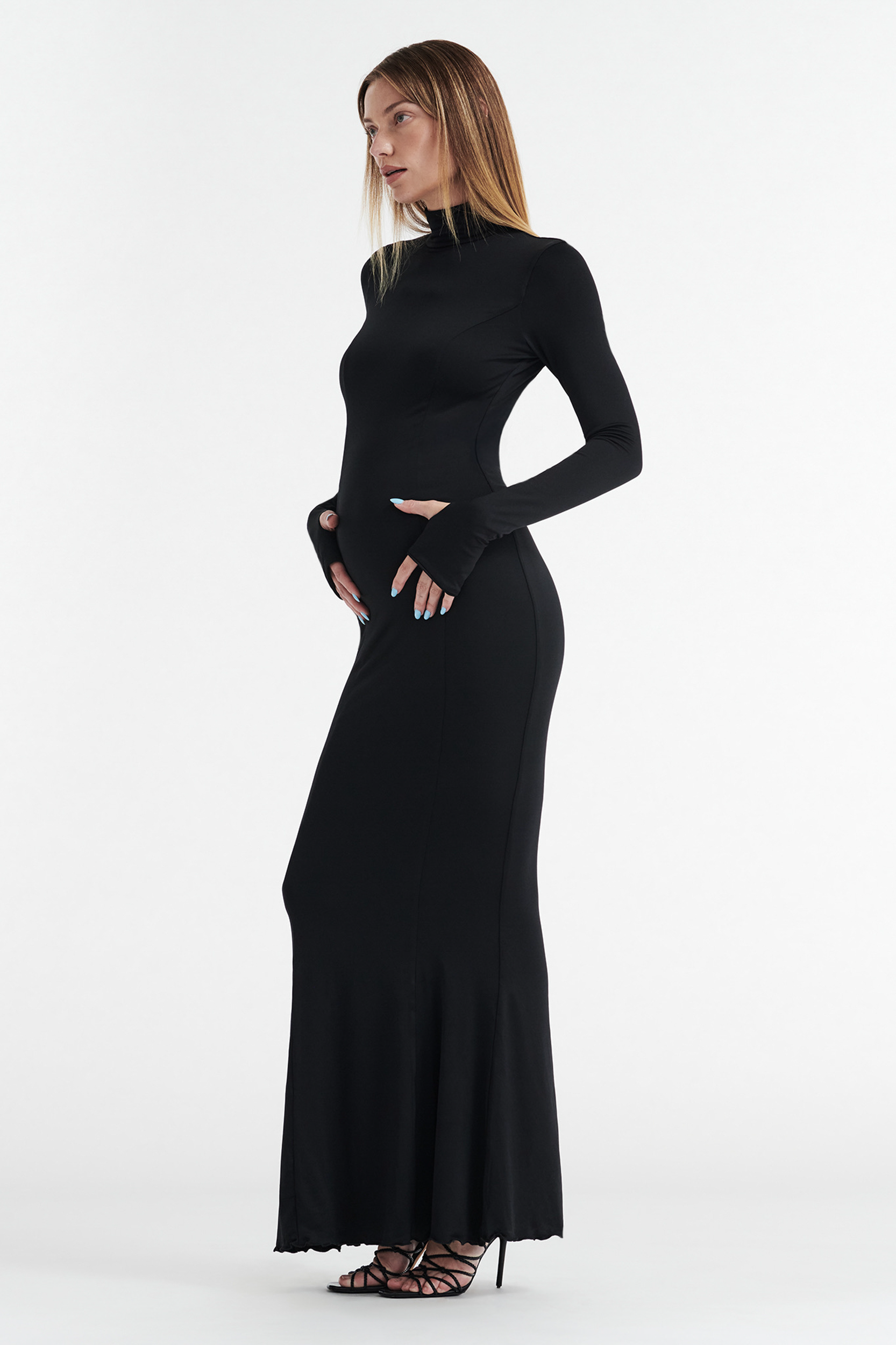 A woman with straight blonde hair models the CDP AUDREY TURTLENECK LONG SLEEVE MAXI DRESS in black, standing in profile against a plain white background with her hands on her hips and wearing black strappy high heels.