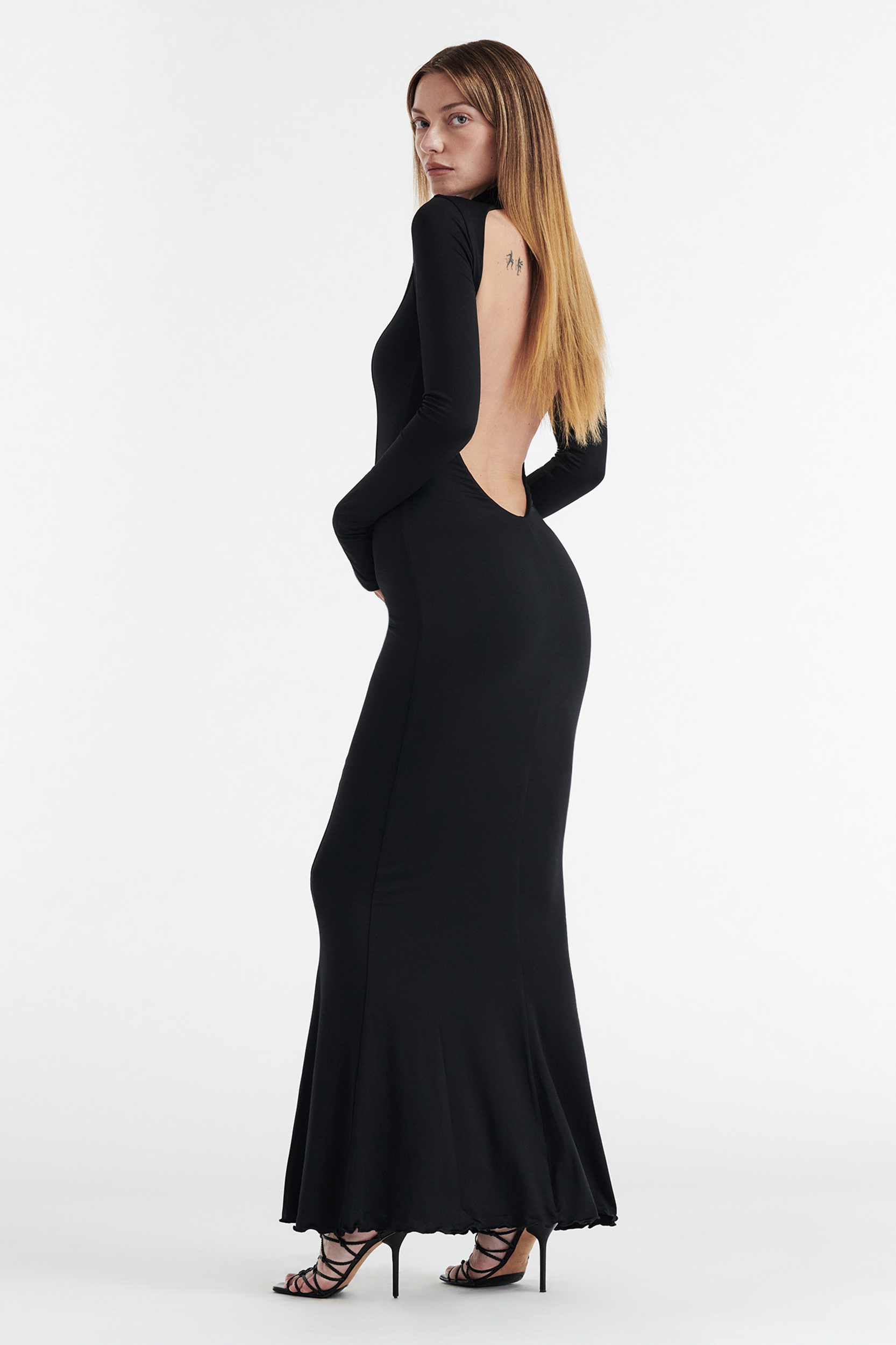 A woman with long straight hair wears the CDP AUDREY Turtleneck Long Sleeve Maxi Dress in black, featuring a dramatic open back and mermaid hem, paired with black strappy heels against a plain white background.