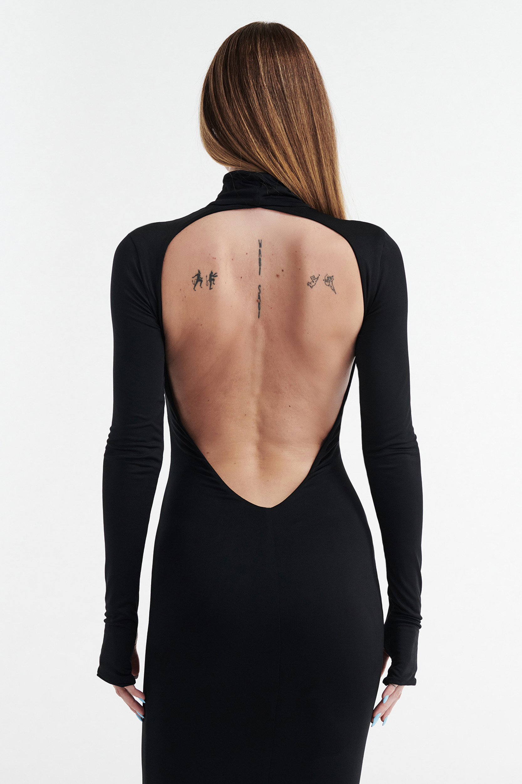 A person with long straight hair models the CDP AUDREY TURTLENECK LONG SLEEVE MAXI DRESS in black, featuring an open back that reveals small tattoos, posed before a plain white background.