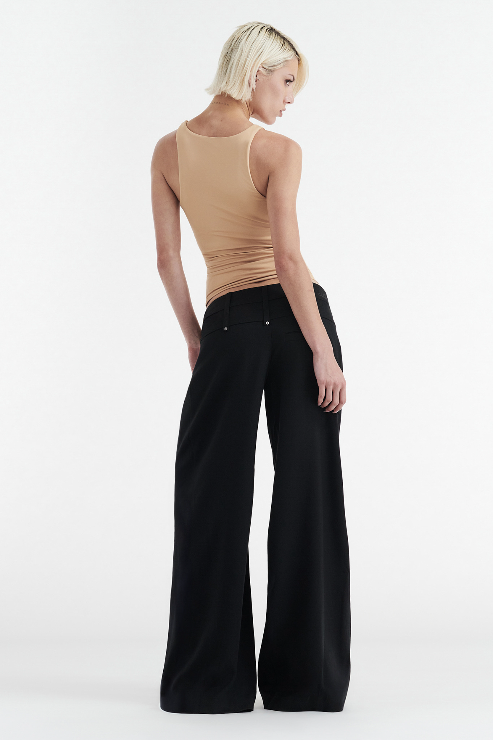 A person with short blonde hair wears the CDP SOPHIE BASIC SLEEVELESS TANK WITH SHELF BRA in tan and high-waisted black wide-leg pants, standing with their back turned and head slightly angled over their shoulder against a plain white background.