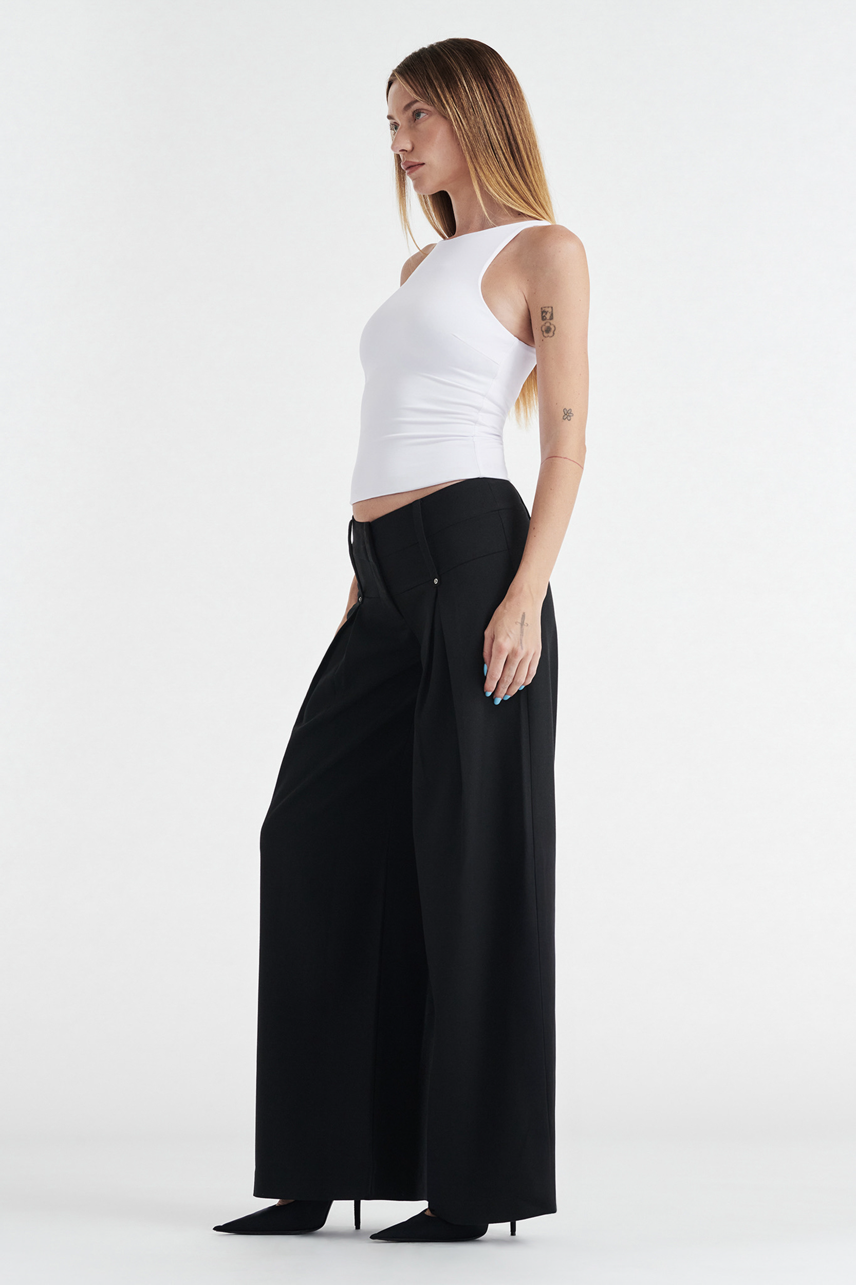 A woman with long straight hair and tattoos on her left arm stands sideways against a plain white background, wearing the CDP SOPHIE BASIC SLEEVELESS TANK WITH SHELF BRA in white, paired with high-waisted wide-leg black pants and black pointed-toe heels.