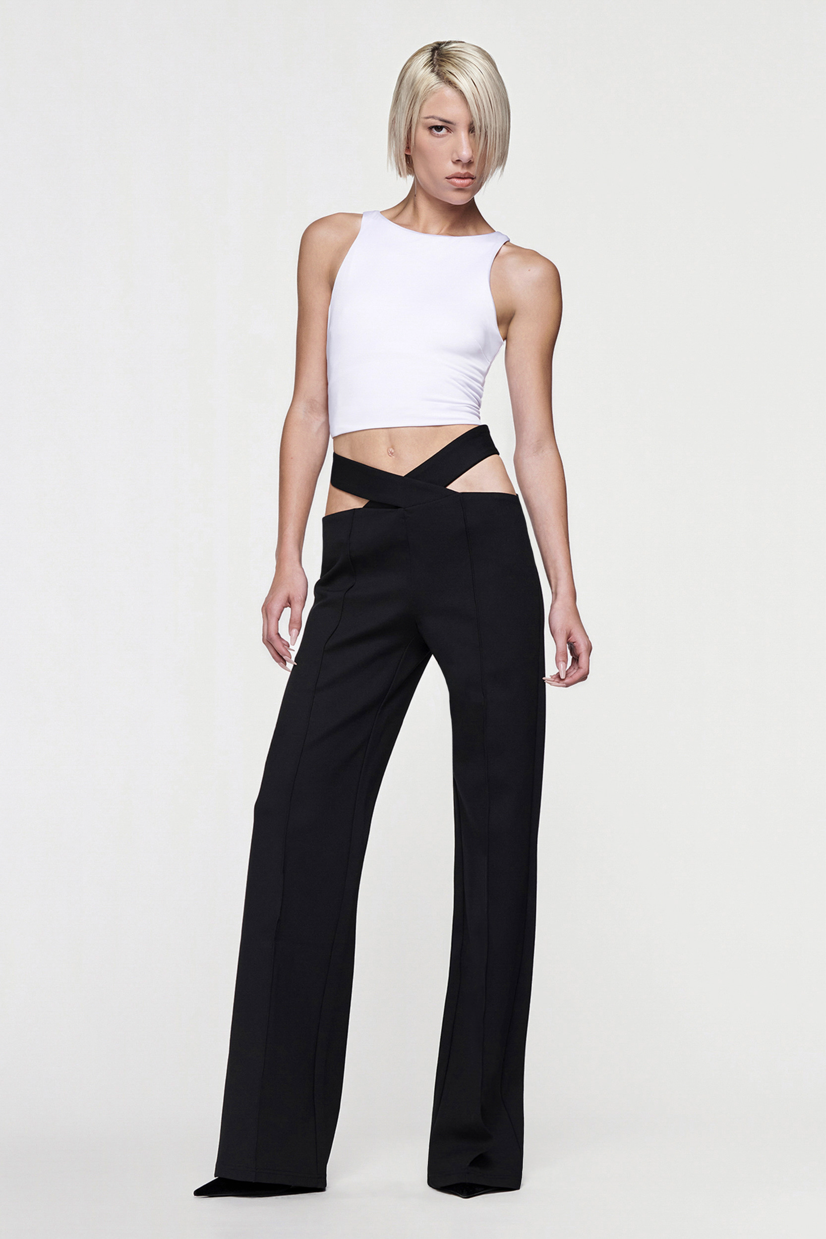 A person with short blonde hair wears a white sleeveless crop top and CDP’s JULIETTE WIDE LEG PANT in black, featuring a high-waisted crisscross waistband, while standing confidently against a plain white background.