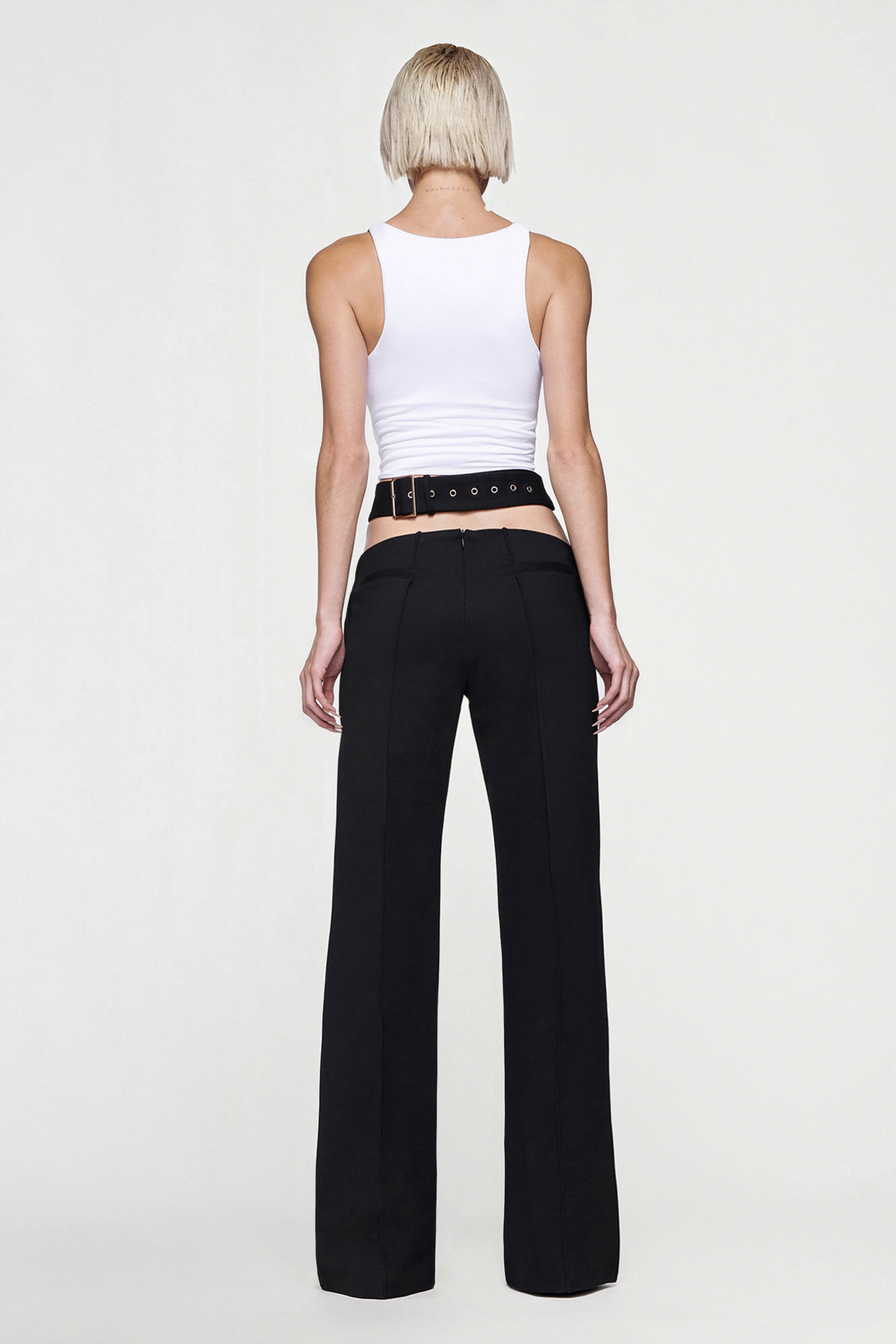 A person with short blond hair faces away, wearing a white sleeveless top and the CDP JULIETTE WIDE LEG PANT in black, styled with a black grommet belt. The background is plain and light-colored.
