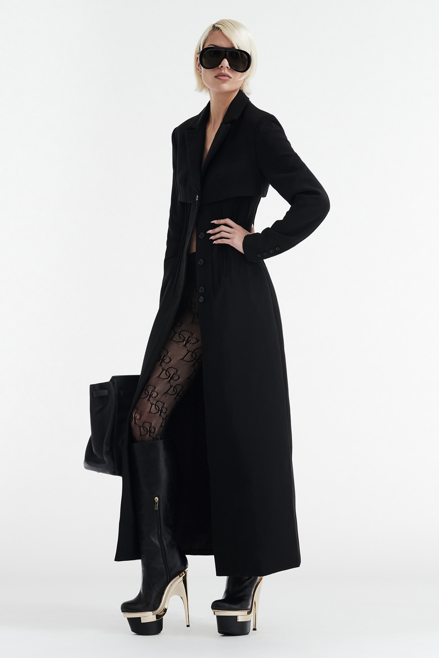 A woman wears the CDP CELINE BUTTON UP TRENCH COAT in black, styled with patterned tights, gold-heeled platform boots, large sunglasses, and a black bag as she poses confidently against a white background.