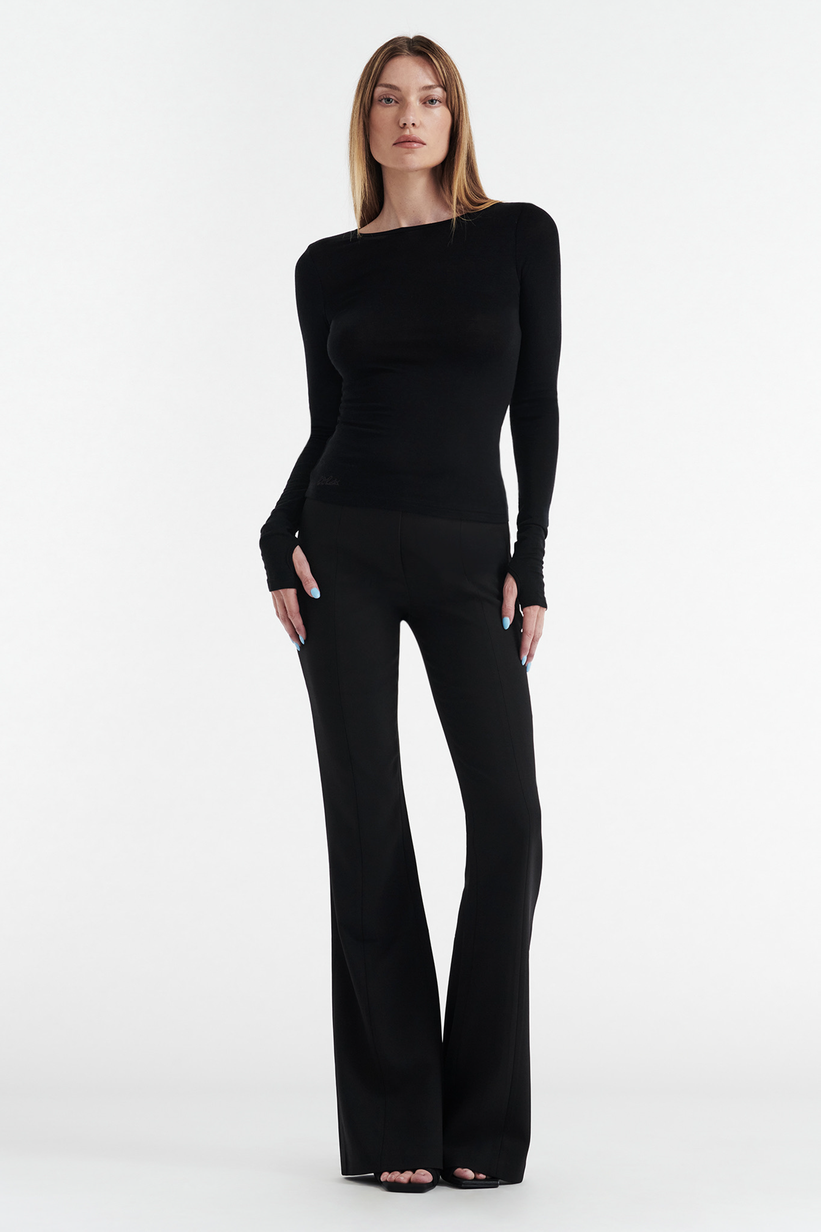 A woman with straight, light brown hair stands against a white background, wearing the CDP CLAUDIA LONG SLEEVE BASIC TOP - BLACK and black flared pants, featuring a fitted silhouette with her hands relaxed by her sides.