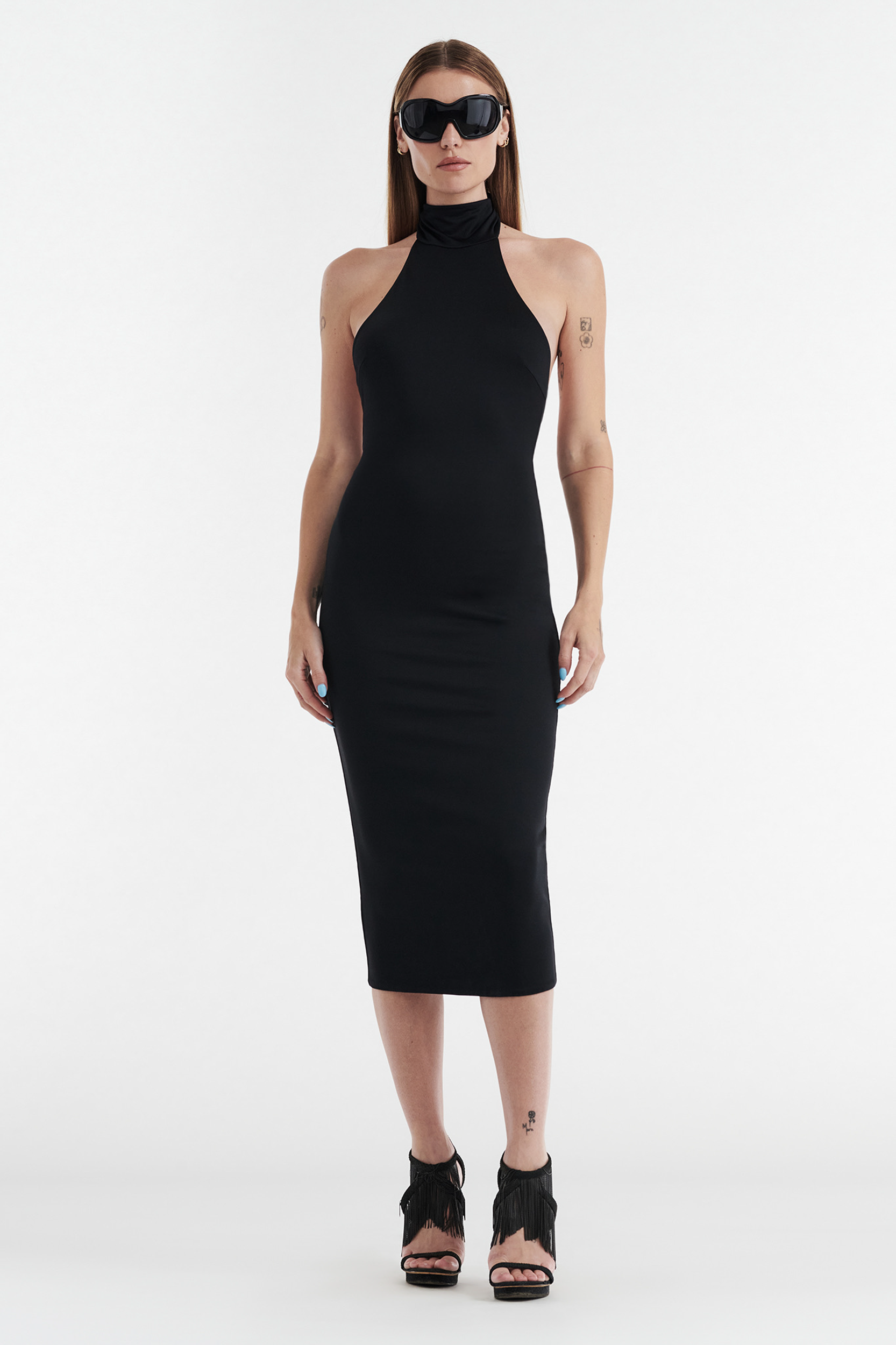 A woman in a sleeveless, fitted CLEO HALTER TIE MIDI DRESS - BLACK by CDP, black fringe high-heeled sandals, and large black sunglasses stands against a white background.