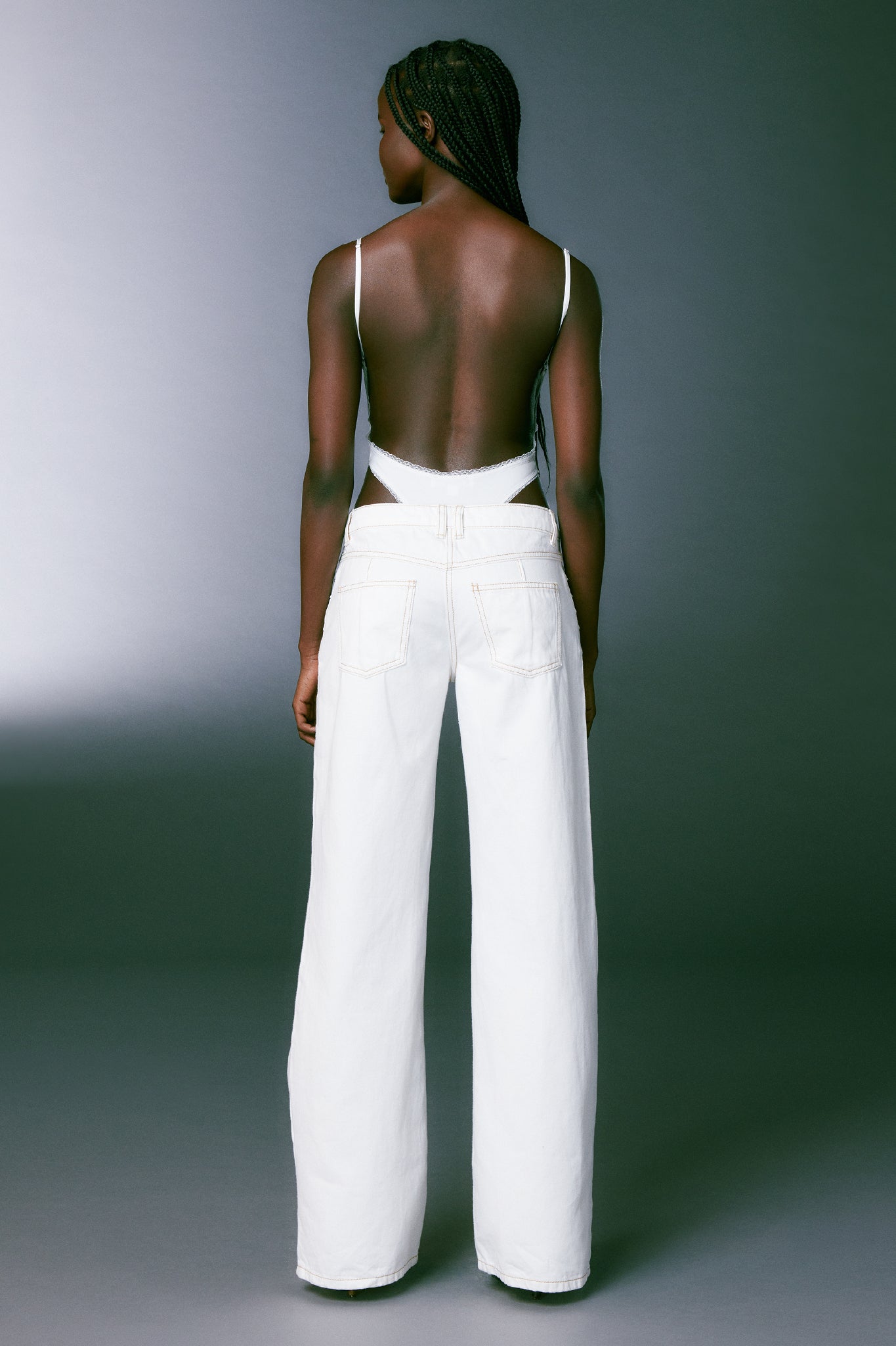 A person with long braided hair is standing with their back to the camera, wearing a backless white top from I.AM.GIA and the DYLAN JEAN - WHITE, high-waisted wide-leg pants by I.AM.GIA. The background features a simple gradient, dark at the edges and lighter near the center.