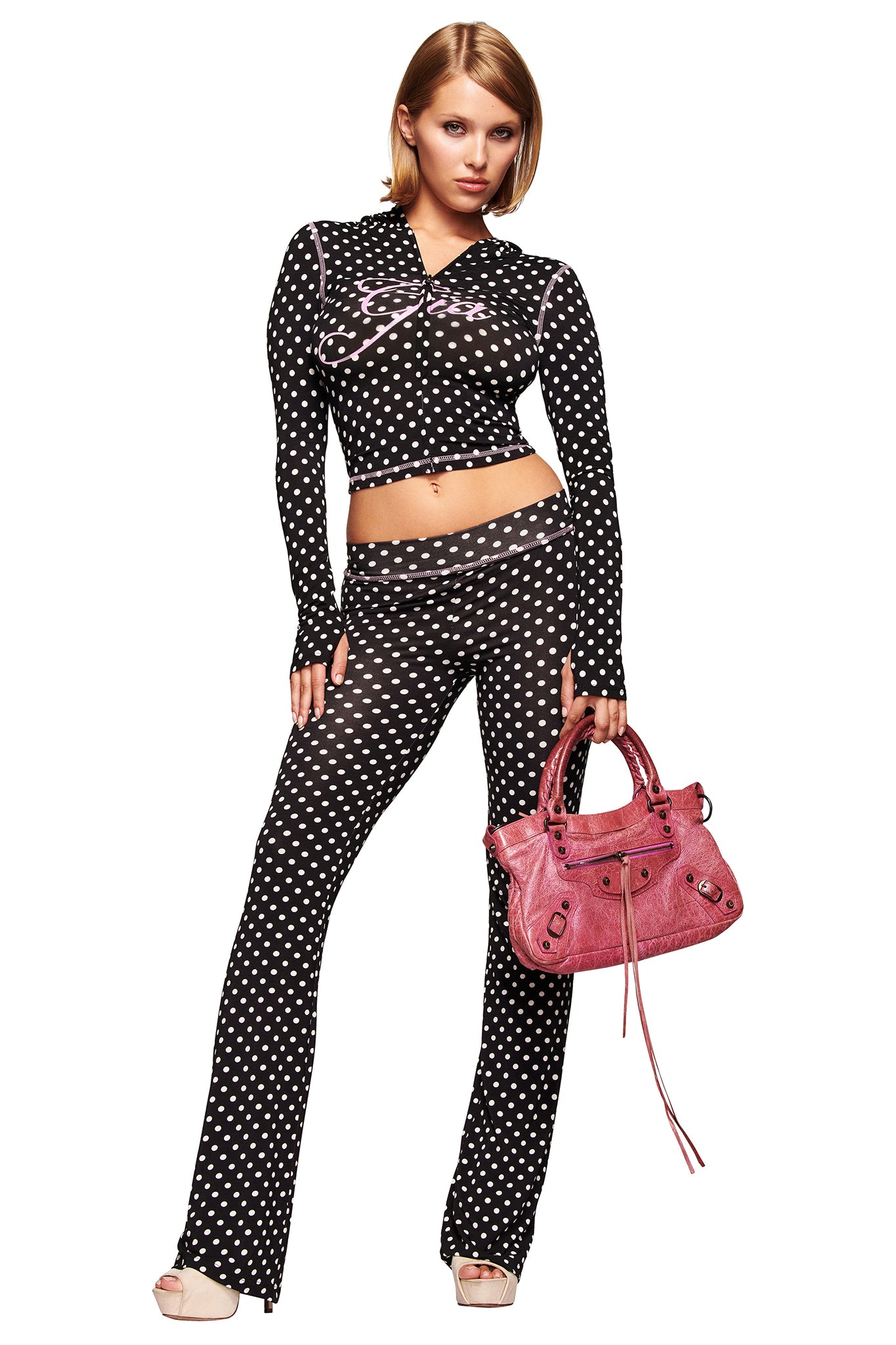 A woman poses confidently against a white background wearing the I.AM.GIA BLARE TRACKPANT in black polka dot, a long-sleeve black crop top, open-toe heels, and holding a pink handbag.