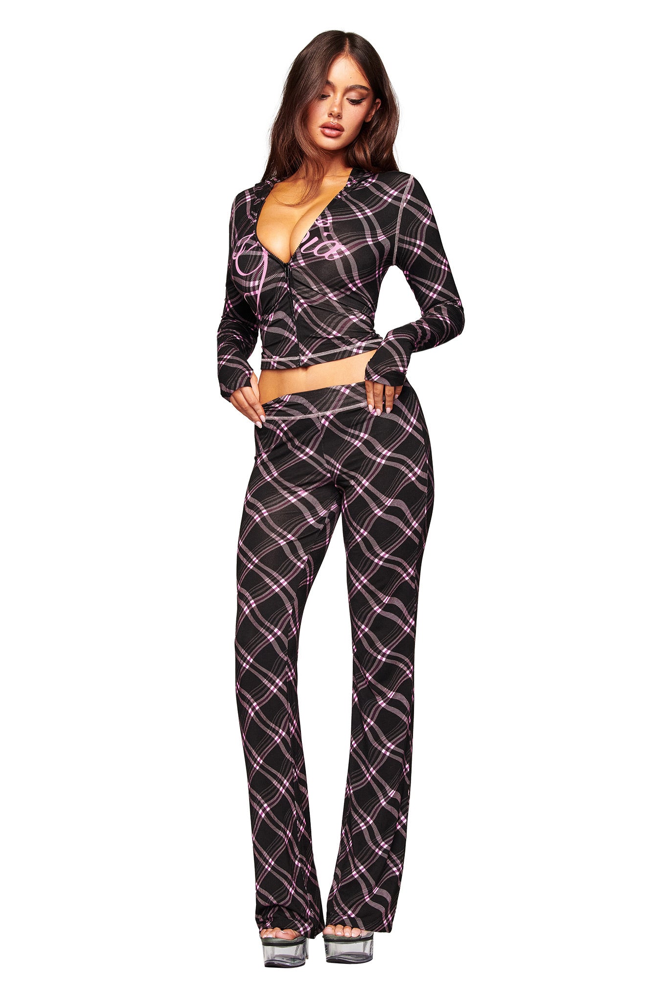 A woman poses against a white background in the I.AM.GIA BLARE TRACKPANT - BLACK WARPED PLAID, paired with a matching long-sleeve deep V-neck crop top and silver platform heels.