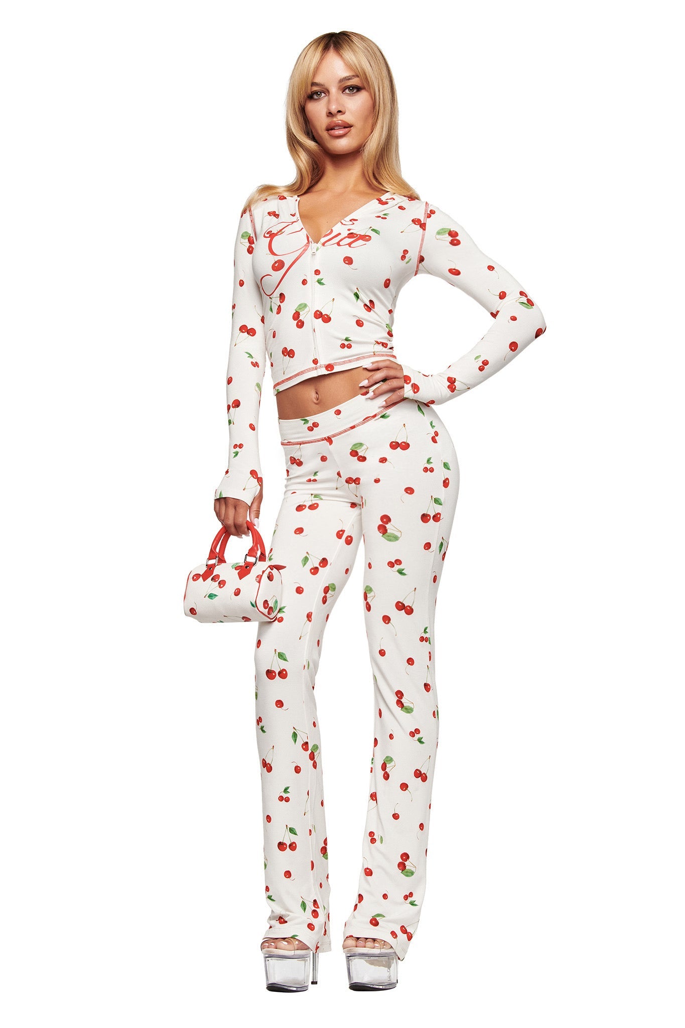 A woman wears the I.AM.GIA BLARE HOODIE in the Cherry on Top print, paired with matching pants and a small handbag, styled with silver platform heels for a bold look.