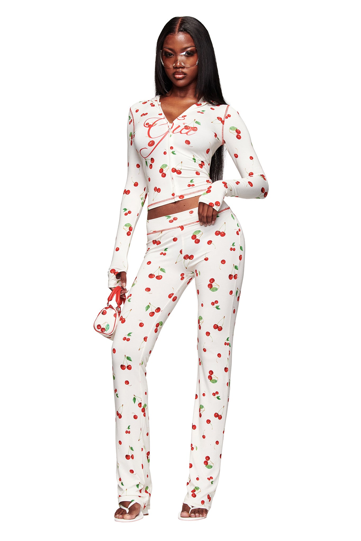 A woman models the I.AM.GIA BLARE TRACKPANT - CHERRY ON TOP with a matching cherry-print purse and long-sleeve white top, confidently posing in heeled sandals against a crisp white background.
