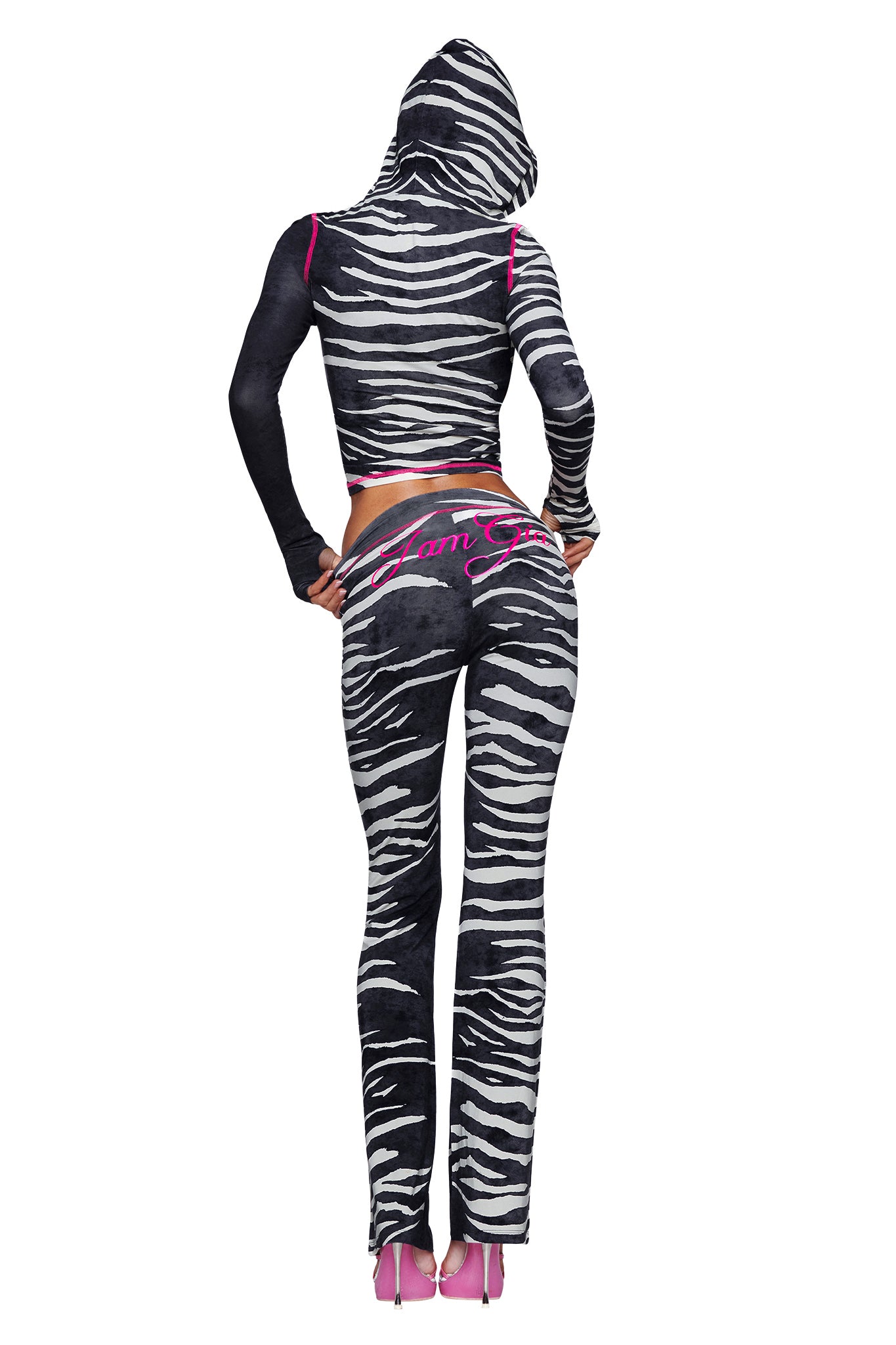 A person stands facing away, wearing the I.AM.GIA BLARE HOODIE - BLACK WILD SIDE—a black and white zebra print hoodie featuring a bold design—paired with matching pants that have pink accents and high heels. The hood is up, and “Glam Girl” is written in pink on the pants.