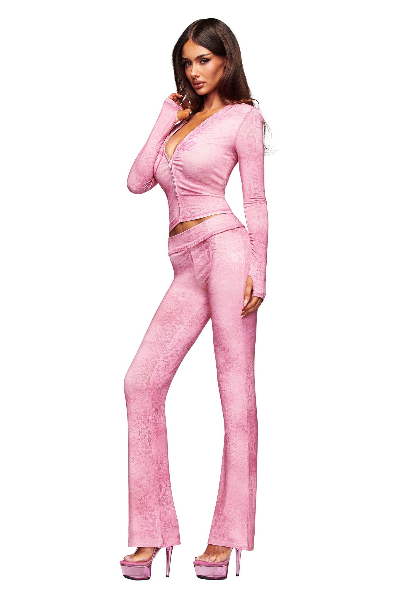 A woman poses confidently in the I.AM.GIA BLARE TRACKPANT - LIGHT PINK SERPENTINE two-piece set with long sleeves and flared legs, styled with matching pink platform heels against a plain white background.