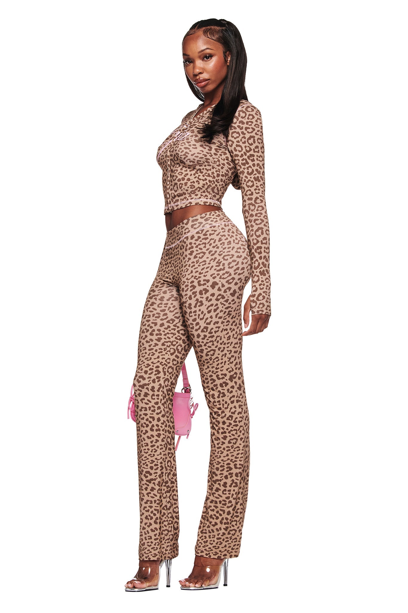 A woman poses against a white background in the I.AM.GIA BLARE TRACKPANT - MOCHA LEOPARD with a matching fitted, long-sleeve leopard print crop top, clear heels, and a small pink handbag.