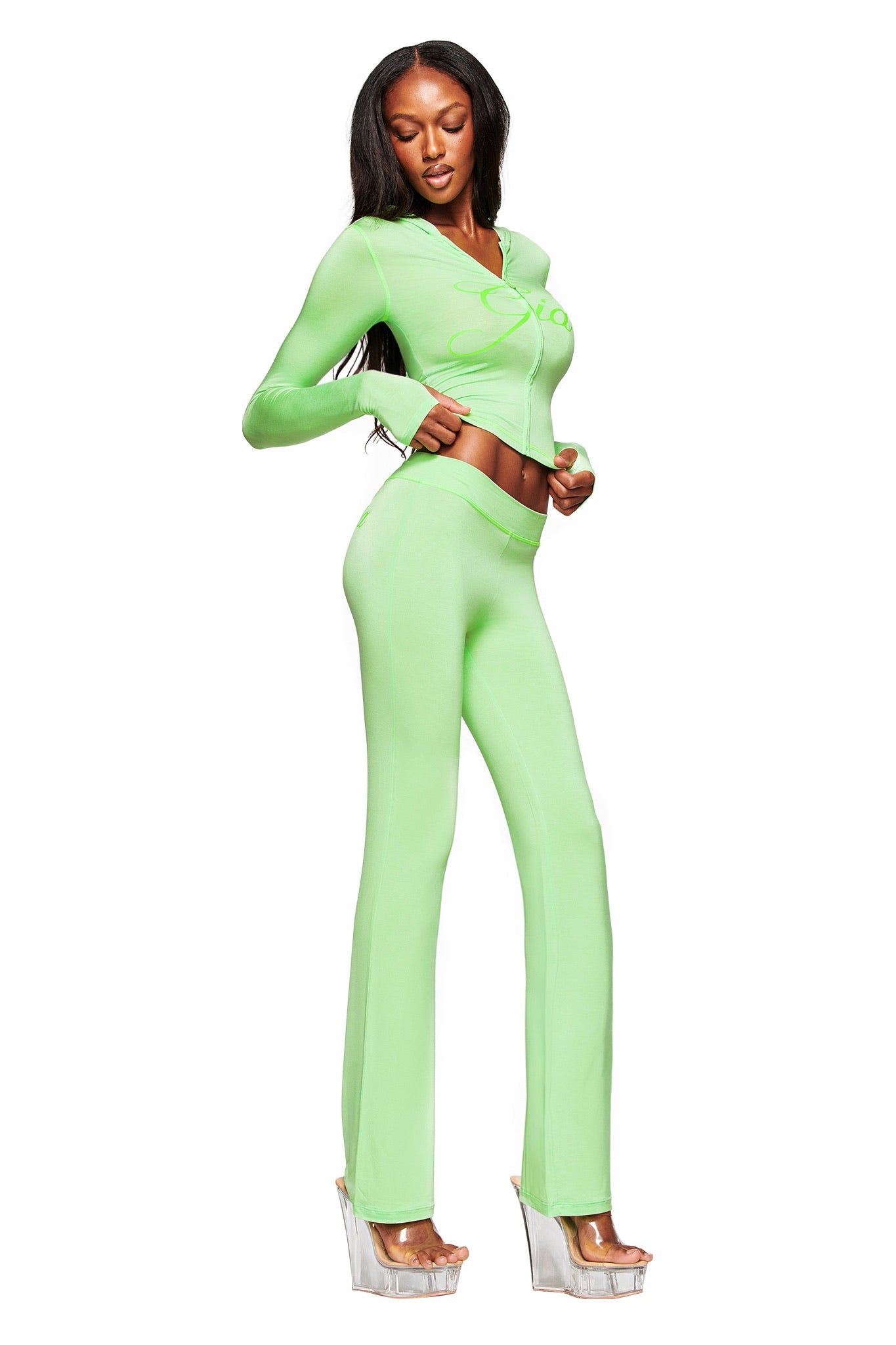 A woman poses in I.AM.GIA’s BLARE TRACKPANT - NEON GREEN, a fitted two-piece set with flared trackpants and long sleeves, styled with clear high platform heels, against a white background.