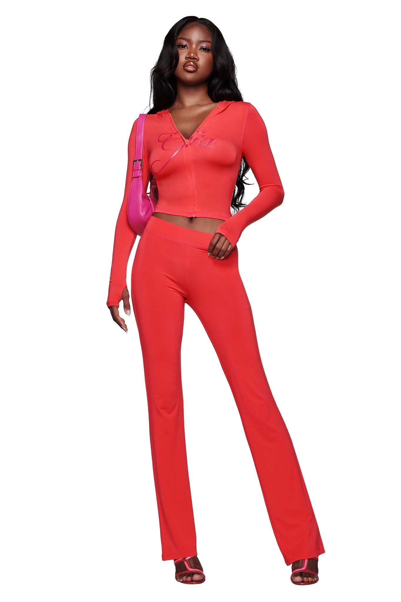 A woman models the I.AM.GIA BLARE TRACKPANT in Papaya, featuring a mid-rise waist and flared leg, styled with a matching fitted red long-sleeve crop top, pink shoulder purse, and red heeled sandals.