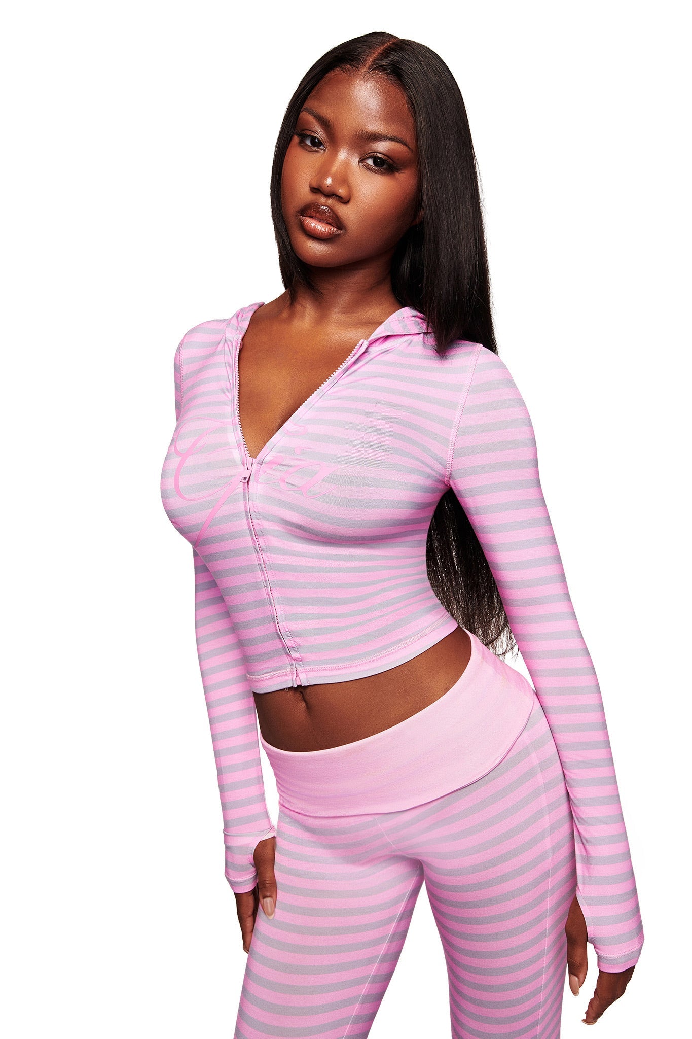 A woman with long straight hair wears the I.AM.GIA BLARE HOODIE - LIGHT PINK STRIPE and matching leggings, featuring horizontal pastel pink and light purple stripes, standing against a white background.